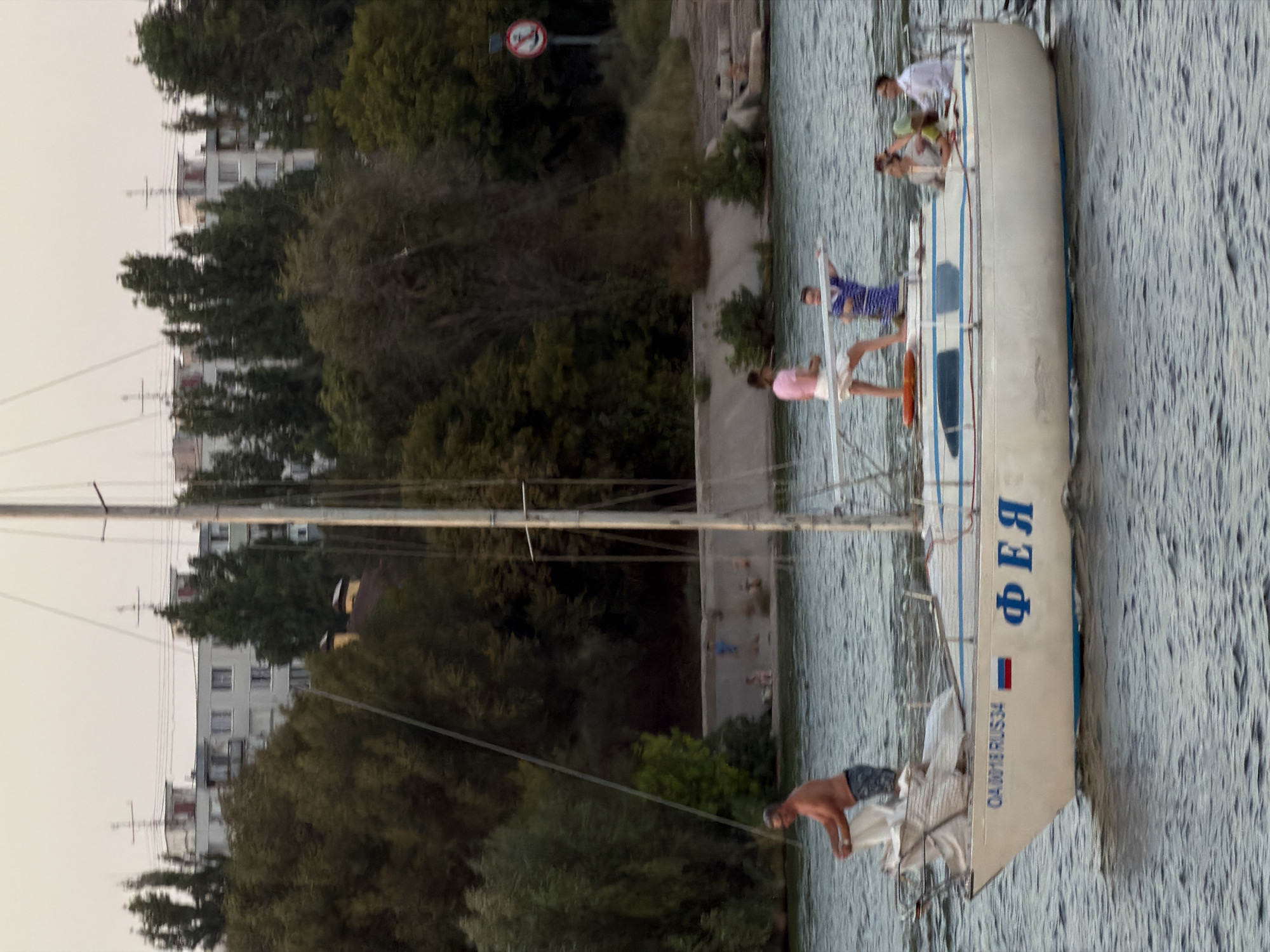 A medium shot captures a sailboat named "ФЕЯ" (Feya) on the water in Kamyshin, Russia, under an overcast sky. The boat is white with blue trim and lettering. Several people are on board. On the front deck, a man in swim trunks is handling some white fabric. On the main deck, a woman in a pink top and white shorts stands near a man in a blue and white striped shirt. Further back, three more individuals are seated. In the background, a concrete embankment is visible, lined with trees and buildings. A sign with an anchor symbol is also present near the embankment. The water is choppy.