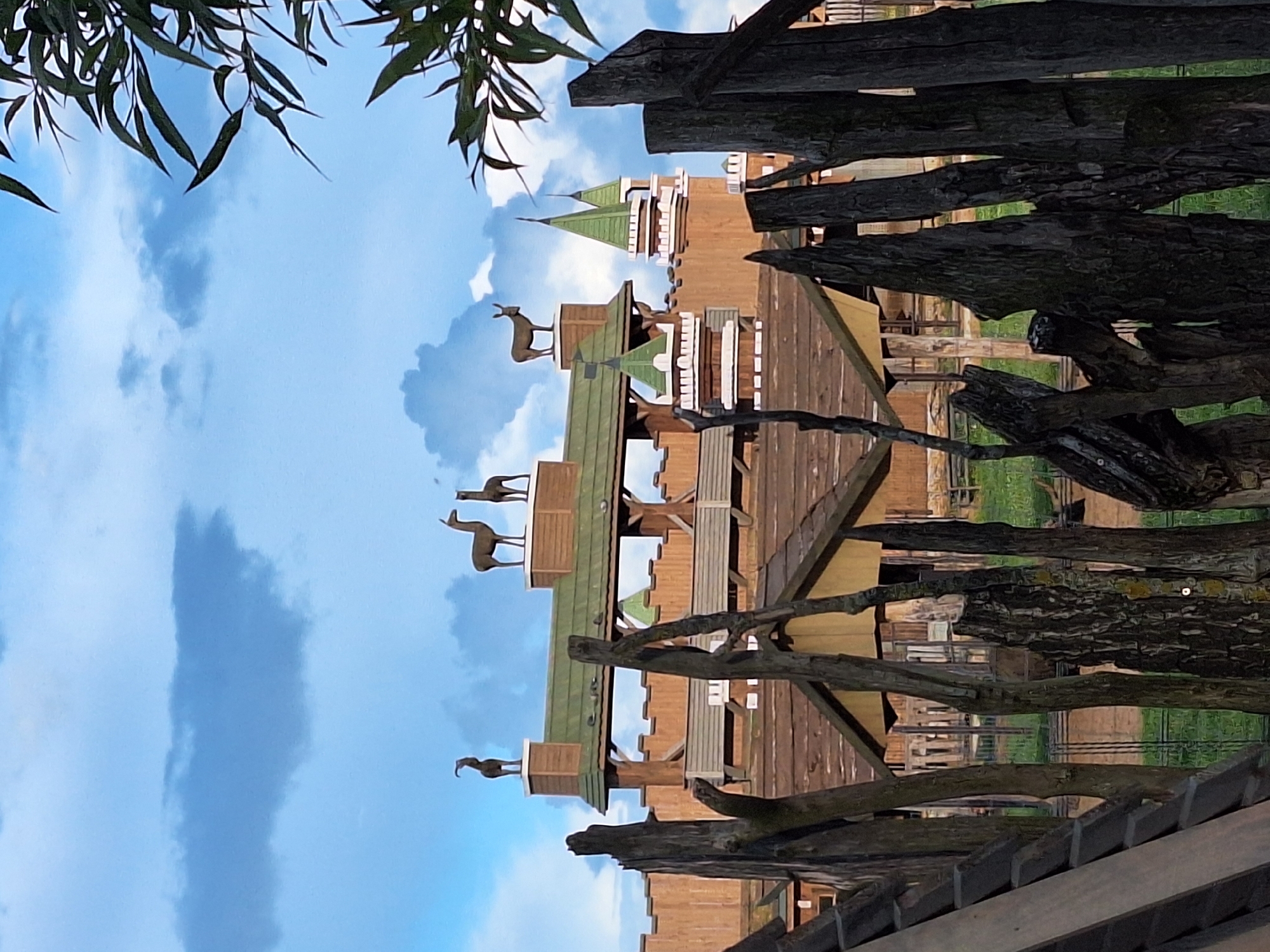The image depicts a partially obscured view of a building complex with a rustic, castle-like architectural style. The structure is made primarily of wood, with turrets topped with green conical roofs. Several animal sculptures, appearing to be deer or similar ungulates, are positioned on top of the highest roof section. In the foreground, a fence constructed of rough, weathered wooden posts and planks partially obstructs the view of the building. The background shows a grassy area and a bright blue sky with scattered white and grey clouds. The scene appears to be outdoors. The specific location in Малахавецкі сельскі Савет, Belarus is not discernible from the visual elements alone. No people are present in the image.