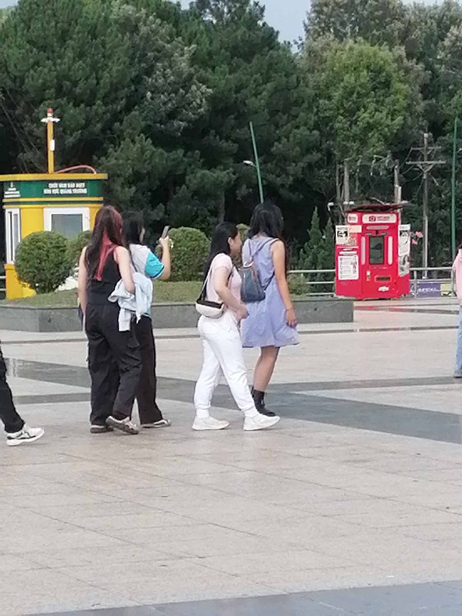 The image captures a street scene in Da Lat, Vietnam. In the foreground, four individuals are walking across a paved area. To the left, two women are walking together, one in a black tank top and black cargo pants, and the other in a white and blue top and dark pants. The woman on the left is carrying a light blue jacket. To their right, a woman in a light blue dress with a black backpack is walking, and further to the right, a person in white pants and a light-colored top is walking away from the camera. In the background, a red vending machine is visible. Behind the vending machine, a yellow and green structure with signage is present. A lush green forest borders the background of the scene.