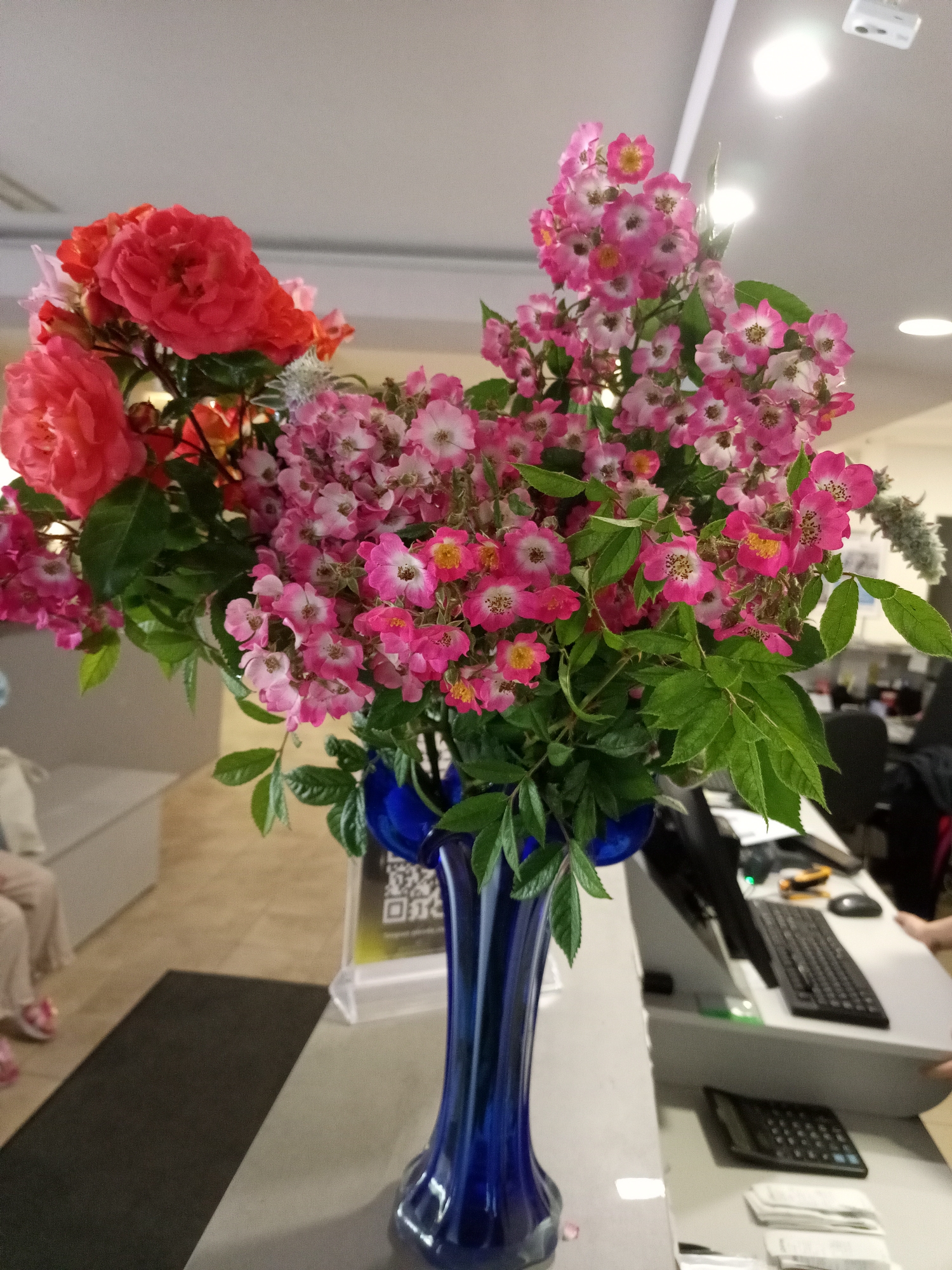 A floral arrangement of pink and red roses is in a blue vase on a counter. The counter has a computer keyboard, mouse, calculator, and a stack of papers. In the background, there are office chairs and desks, suggesting an office environment. A person wearing light-colored pants and pink shoes is partially visible on the left side of the image, sitting on a bench. The scene appears to be indoors.