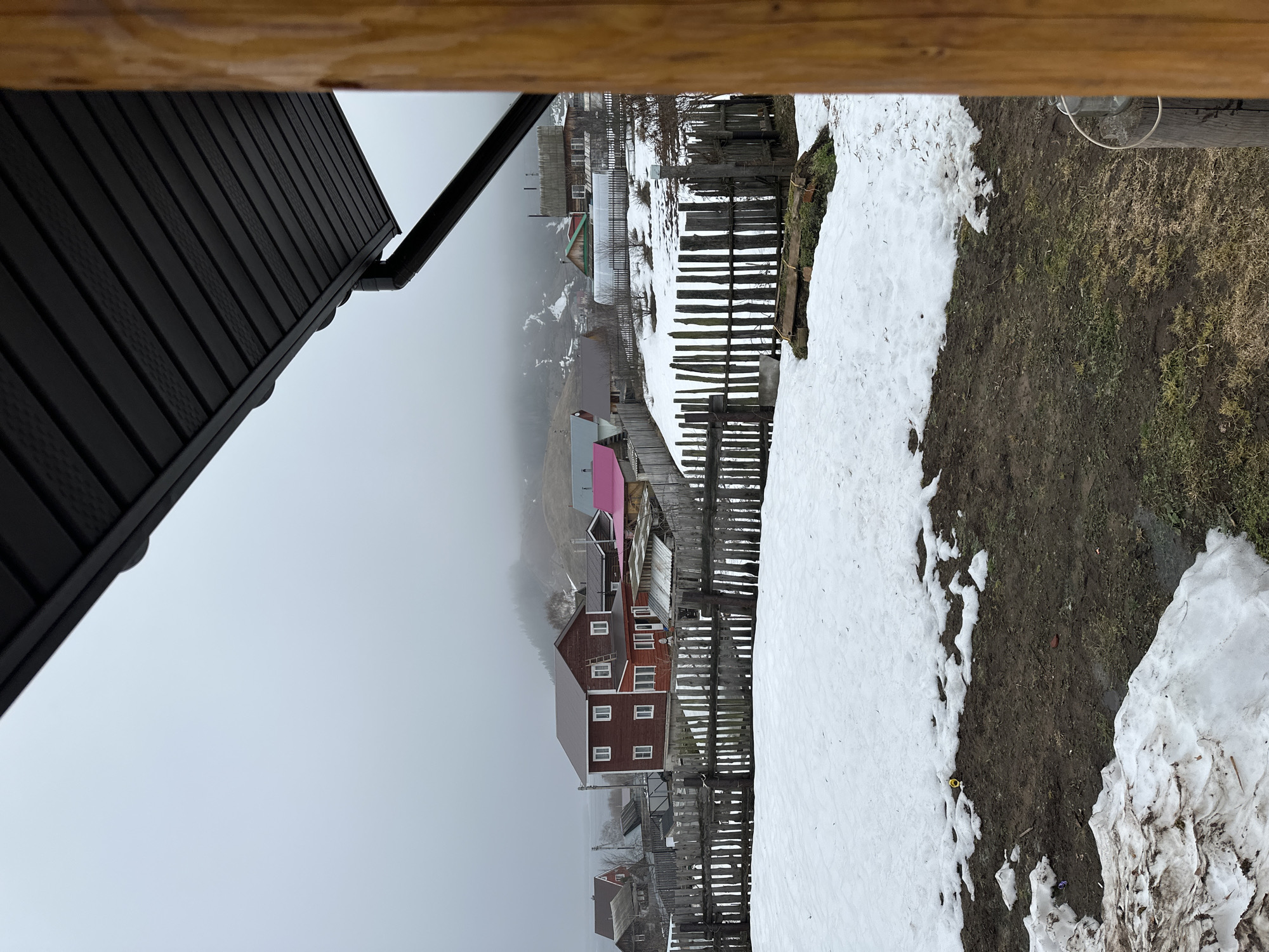 The photograph depicts a wintery scene in Ольховка, Russia. The foreground shows a dirt path with patches of melting snow alongside a wooden fence. In the middle distance, there are several houses. One is a multi-story house with a maroon exterior and white-framed windows. Another building has a pink roof. The background features a foggy hillside. An overhanging dark-colored roof is visible in the upper left corner of the image. A weathered wooden beam is in the top center of the image.