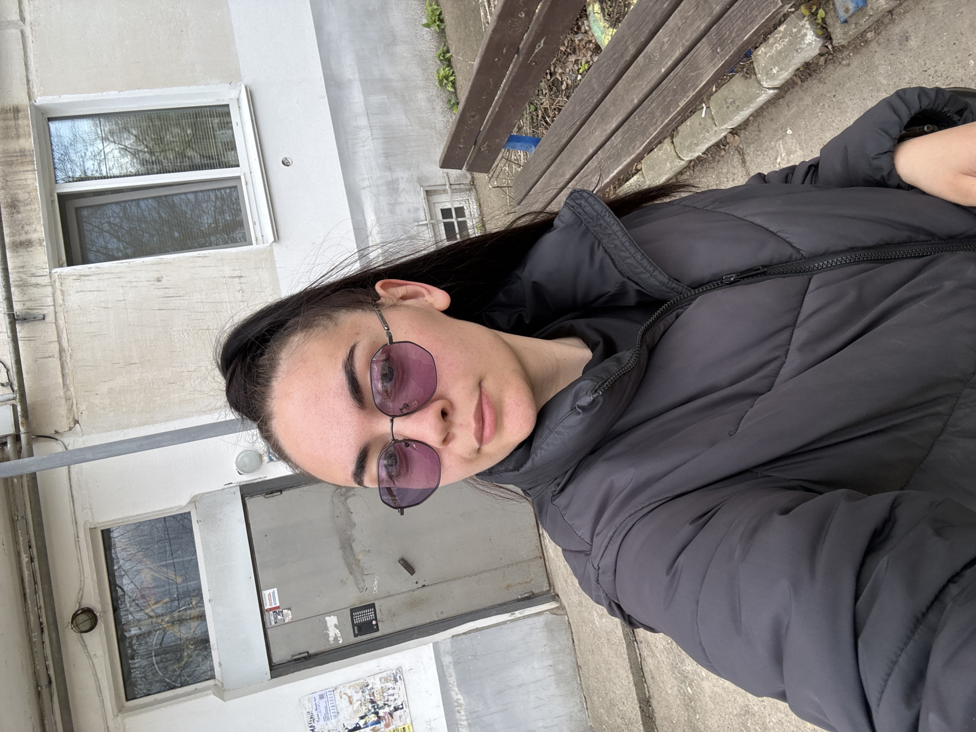 A young woman is seated on a wooden bench outside a multi-story building in Chaikovsky, Russia. The building is constructed of painted concrete, featuring rectangular windows and a metal door. The woman wears a black puffer jacket and round sunglasses with a purple tint. The background includes concrete sidewalks and sparse vegetation. The overall atmosphere suggests a casual, everyday setting.
