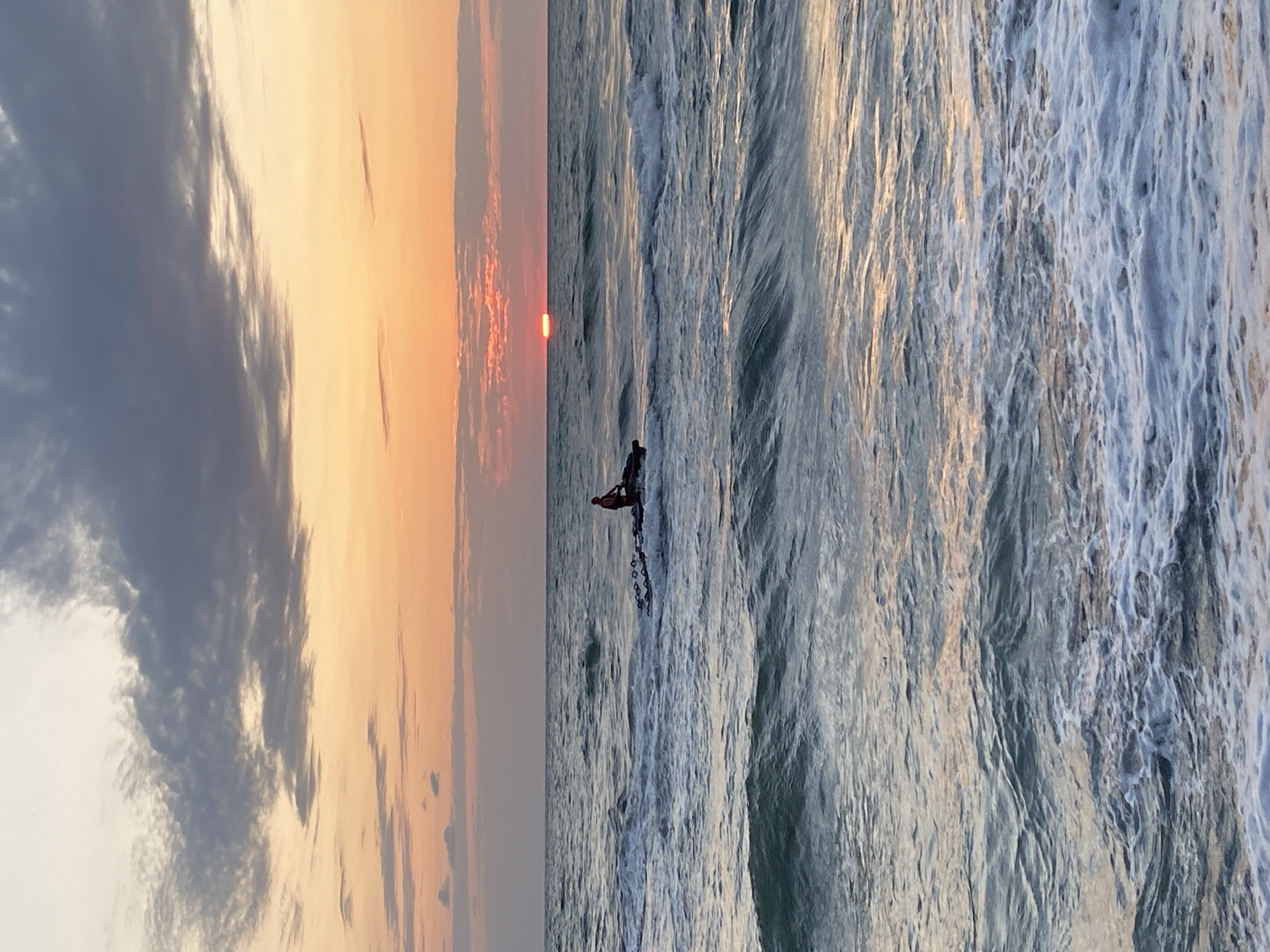 A man in a red top is riding a jet ski in the sea off the coast of Ureki, Georgia. The sun is setting on the horizon, casting an orange glow over the sky and water. The waves are moderate, with some white foam visible. The sky is partly cloudy, with dark clouds in the upper left corner and lighter clouds closer to the horizon.
