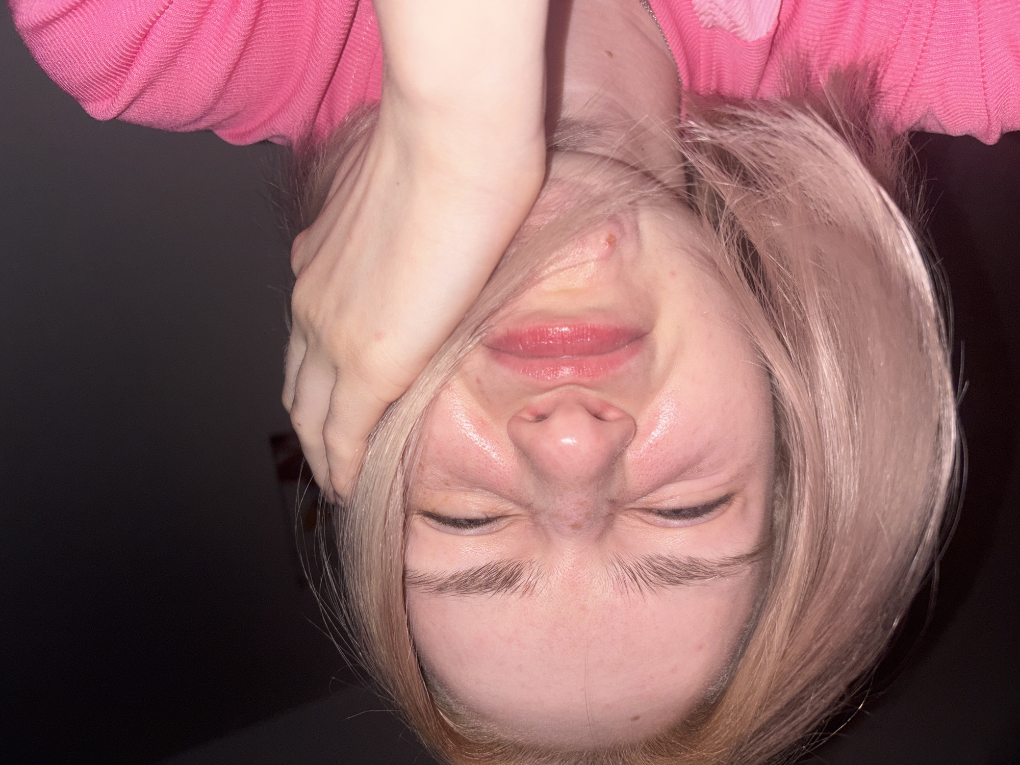 The image presents an upside-down, close-up portrait of a young individual, likely female, situated in Vsevolozhsk, Russia. The person has fair skin and light, possibly blonde or light brown hair, which frames their face. They are wearing a vibrant pink, ribbed long-sleeved top or sweater.
The subject's face displays a strong, expressive grimace: their eyes are tightly shut and squinted, the nose is scrunched, and the lips are slightly parted in a contorted expression. One hand, with fair skin, is positioned on the upper left side of their face, partially covering the hair and forehead. The skin on the forehead and nose appears somewhat shiny, indicating a slight sheen or oiliness. Several minor blemishes are visible on the forehead, bridge of the nose, and chin area.
The background is uniformly dark and featureless, suggesting an indoor setting with low ambient lighting. No specific objects or textual elements are discernible in the background. The time of day cannot be determined from the image, but the lighting suggests it could be evening or night, or a dimly lit interior. The overall impression is one of intense emotion, possibly playful discomfort, strain, or a deliberately exaggerated facial expression.