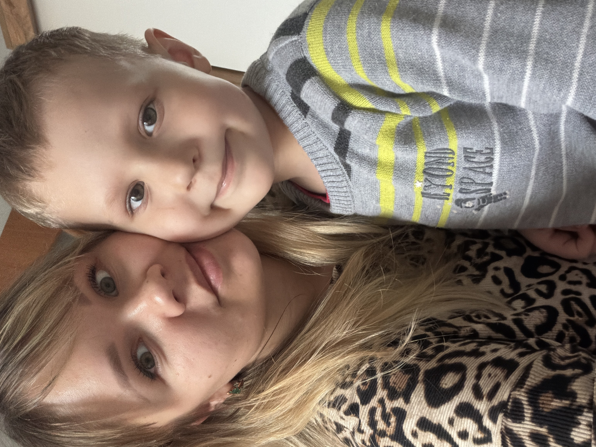A close-up selfie shows a woman and a young boy lying close together. The woman has blonde hair and is wearing a leopard-print top. The boy has short brown hair and is wearing a gray striped sweater with yellow accents and the words "Beyond Space" on it. Their faces are near each other, and they are both looking at the camera. The location is Unknown, Unknown.