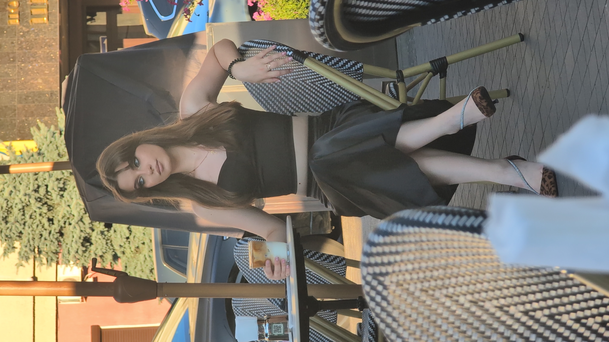 A young woman with long, brown hair is sitting at an outdoor table, holding a drink with a straw. She is wearing a black top and black wide-leg pants. Her shoes are open-toed with a leopard print design and embellished straps. The table and chairs appear to be part of a cafe or restaurant setting, with black and white patterned seating. In the background, there are trees and a building. The lighting suggests it is daytime, possibly late afternoon due to the warm, golden hue. The woman's gaze is directed towards the camera with a neutral expression. She is wearing jewelry, including a bracelet and a necklace. There is a QR code visible on the table.