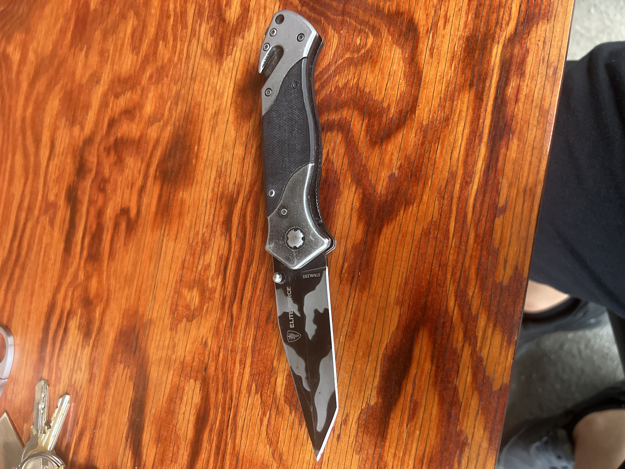 A folding tactical knife with an open blade is positioned diagonally on a polished, reddish-brown wooden table. The knife features a dark camouflage-patterned tanto-style blade with "ELITE FORCE" and a shield-like logo engraved near the hilt, and "STAINLESS" oriented vertically. The handle is composed of metallic silver-grey components and dark, textured grip inserts, secured by multiple screws. The pommel includes an integrated seatbelt cutter/hook and a pointed glass breaker. In the upper left corner, a set of keys on a keyring, made of silver-colored metal, are partially visible. The wooden table exhibits prominent grain patterns and reflects overhead lighting. Below the table level, in the lower frame, parts of a person's legs and feet are visible, one wearing a dark shoe and the other partially covered by dark fabric.