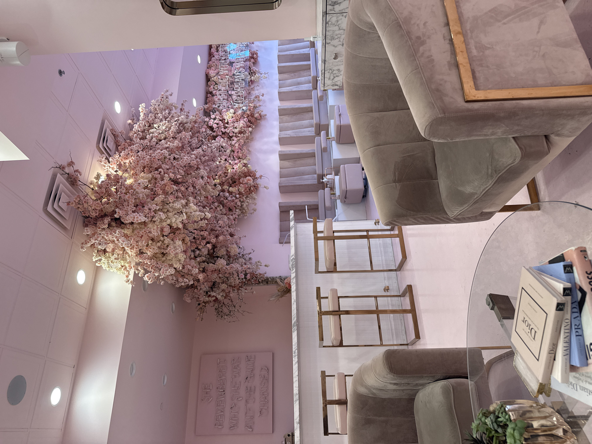 Here is a description of the image:
**Scene Overview:** The image captures the interior of a salon or high-end beauty parlor. The dominant colors are soft pinks, creams, and muted golds, creating a luxurious and feminine atmosphere. The decor incorporates elements of nature and elegance.
**Objects and Subjects:**
* **Seating:** There are multiple plush, gray-beige armchairs arranged around a round glass table. The seating appears comfortable and designed for extended relaxation or treatments.
* **Tables:** A glass-topped table sits nearby, holding a stack of fashion books including Dior, Valentino, and Prada. A small green plant serves as a decorative accent on the table.
* **Manicure Stations:** Multiple manicure stations are visible, each equipped with a chair and setup for nail treatments.
* **Floral Arrangement:** A large, artificial cherry blossom tree or wall installation dominates the left side of the frame, adding a whimsical and romantic touch.
* **Wall Decor:** A pink neon sign with the text "GIRRREUL, YOU BETTER MAKE YOUR DREAMS WILEY" is mounted on the wall, near a painted sign with the text "SHE REMEMBERED WHO SHE WAS AND THE SAME CHANGED."
**Environment:** The scene is well-lit and appears meticulously designed. The space is clean and modern, with a focus on aesthetics and customer experience.
**Location:** Given that the city is Glendale, United States of America, the location is likely a high-end beauty parlor or salon in a retail or commercial area.