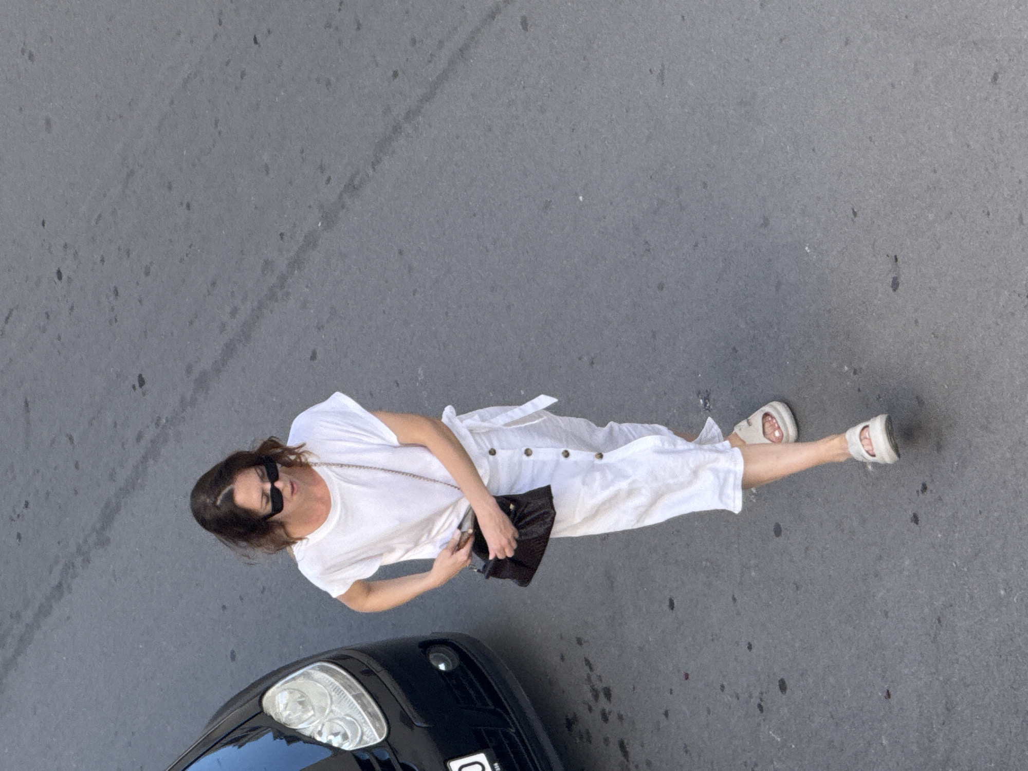 A medium shot from a high angle shows a woman with brown hair walking on a gray asphalt street in Budva, Montenegro. She is wearing a white t-shirt, a white maxi skirt with buttons down the front, and white slip-on sandals. She has sunglasses on and is holding a dark brown clutch bag, with her left hand reaching into it. A portion of the front of a black car is visible to the left of the woman, with its headlight and part of the license plate showing. The street appears to be damp in some areas and has some dark spots.