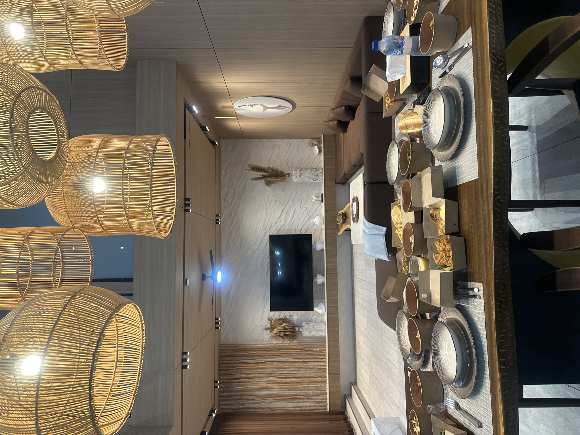 A high-angle interior shot shows a dining area and living room in a modern, possibly coastal-inspired, residence in Kuta Selatan, Indonesia.
The dining area, positioned towards the right, features a long, wooden dining table set with tableware including plates, bowls, cutlery, and takeout containers, indicating a recent meal. The table is partially obscured by black dining chairs with dark wood legs.
Adjacent to the dining area is a living room with a brown sectional sofa. A light blue towel or blanket is placed casually on the couch.
In the background is a textured wall resembling marble or a natural stone pattern. A black flat-screen TV is mounted on the wall, centered in the image. Below the TV, there's a decorative arrangement featuring dry grasses or plants and a horizontally-lined wooden panel.
The lighting is dominated by several large, woven rattan pendant lamps hanging from the ceiling, providing a warm, ambient glow. The scene also includes a ceiling fan.
The overall setting suggests a comfortable, stylish living space.