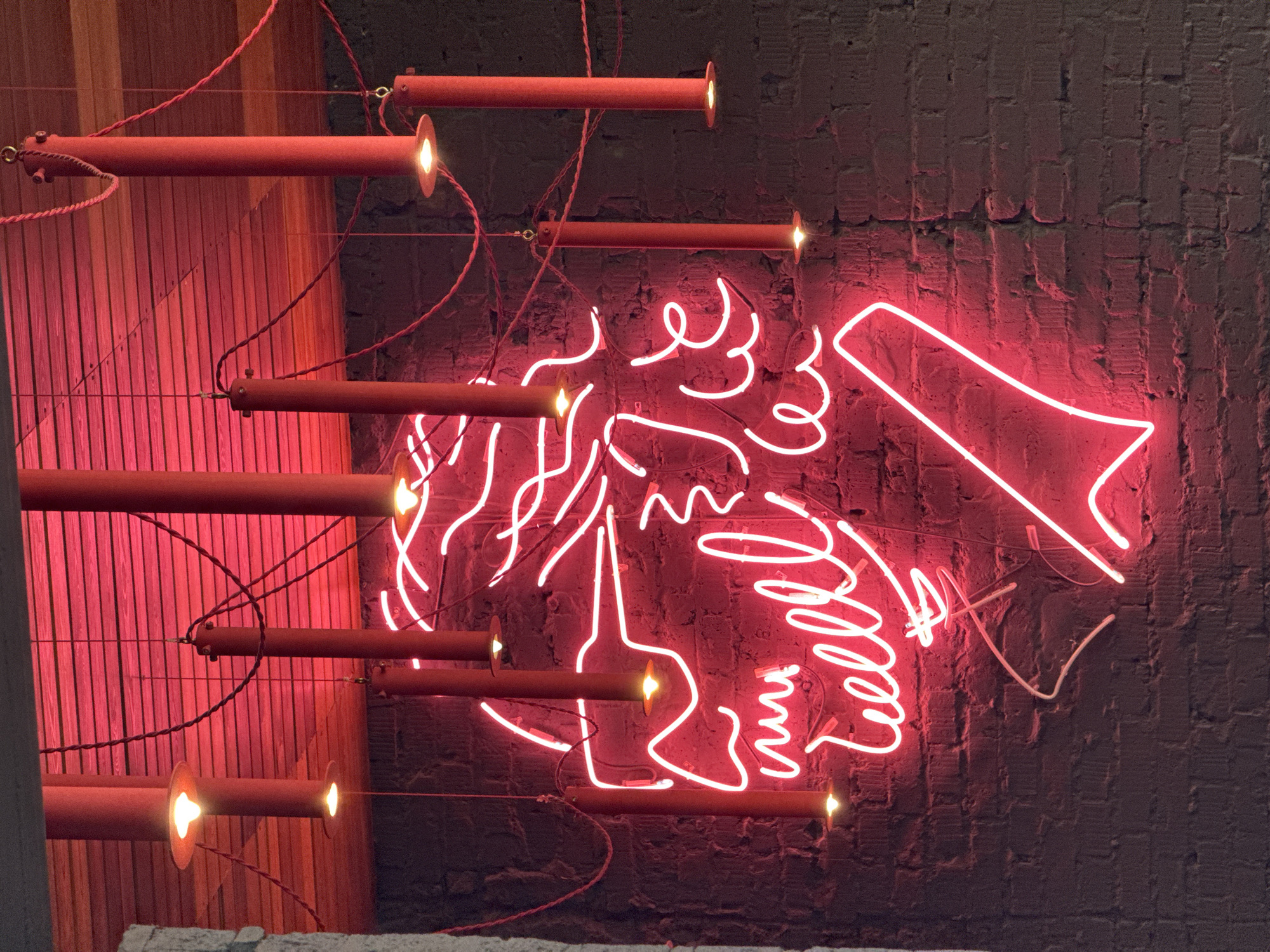 The image presents a vibrant, dimly lit interior space characterized by modern artistic lighting. A prominent feature is a glowing red neon sign against a dark, textured brick wall. The neon sign depicts a stylized profile of a head or face, possibly a woman, facing towards the right. Her "hair" or surrounding elements are rendered as dynamic, squiggly red lines that traverse a significant portion of the frame. To the right of the face, the neon forms the distinct shape of a megaphone or speaker, suggesting themes of communication or announcement. Below the face, some of the squiggly lines appear to form highly stylized, possibly illegible cursive text, vaguely resembling "Hello."
In the foreground, several red, tubular light fixtures are suspended, their ends emitting warm light. These fixtures are interconnected by red braided electrical wires that weave through the scene, contributing to an industrial-chic aesthetic. The background behind the neon sign is a dark brick wall, while to the left, a vertical wooden slatted wall is visible, illuminated with a warm red glow.
The scene appears to be set at night or in an artificially lit indoor environment, such as a bar, restaurant, or creative space. The dominant colors are striking reds and pinks from the neon and accent lighting, contrasting with the dark, muted tones of the walls. There are no overt visible textual cues or specific geographical identifiers within the image itself.