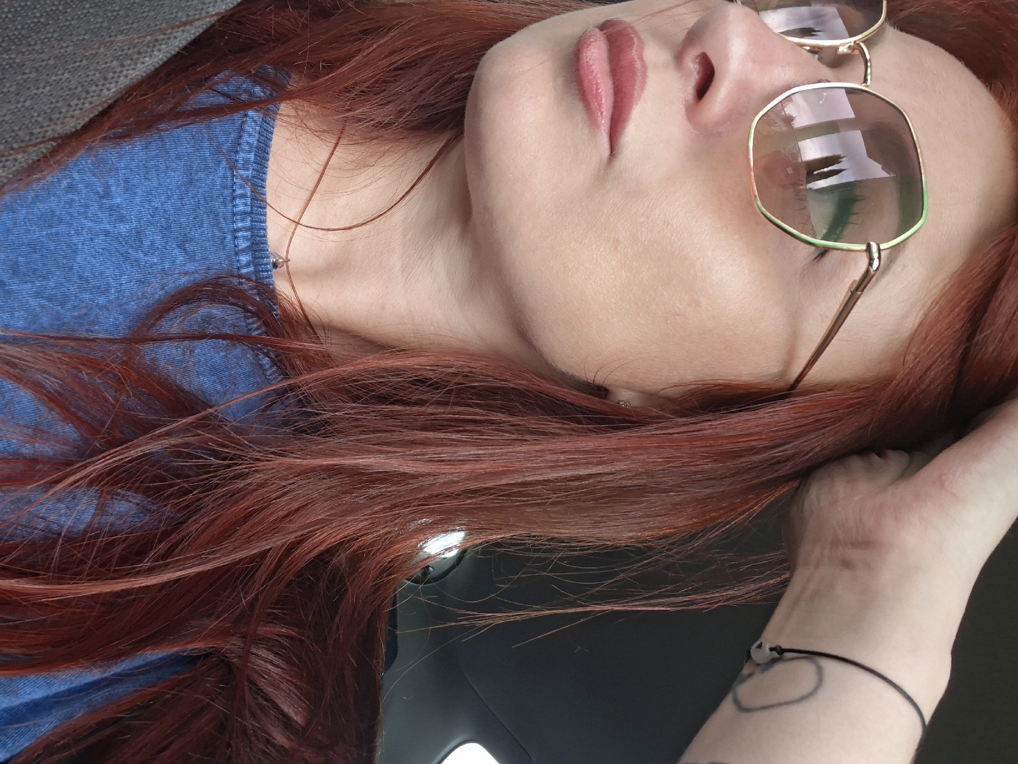 A medium close-up shot shows the upper body of a person with auburn hair. They are wearing sunglasses with an octagonal gold frame and green accents on the upper rim. The person has fair skin and is wearing a blue denim-like shirt. A thin silver chain with a small pendant is visible around their neck. The background is dark and blurred, suggesting an indoor setting such as a car interior. The person's right hand is raised, with fingers touching their hair. A black string bracelet with a small pale bead is visible on their wrist, along with a faint tattoo on their forearm. The image is a close-up and does not provide any details about the city of Myadzyel, Belarus.