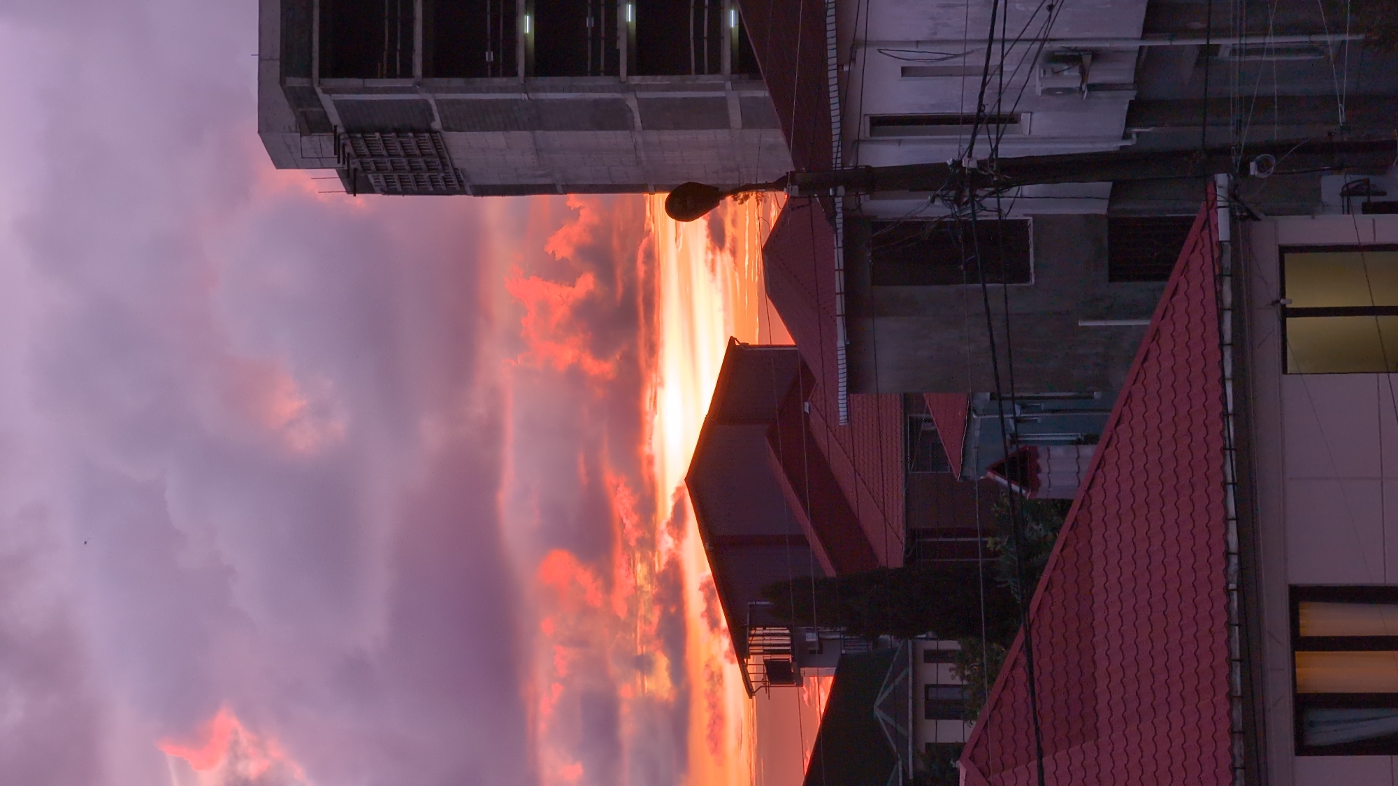 The image depicts a sunset over a city, likely Kobuleti, Georgia. Buildings of varying heights and construction are visible. The sunset creates vibrant orange and red hues in the sky, contrasting with the purple and gray clouds. Electrical wires and a lamppost are in the frame. The image is rotated 90 degrees counter-clockwise.