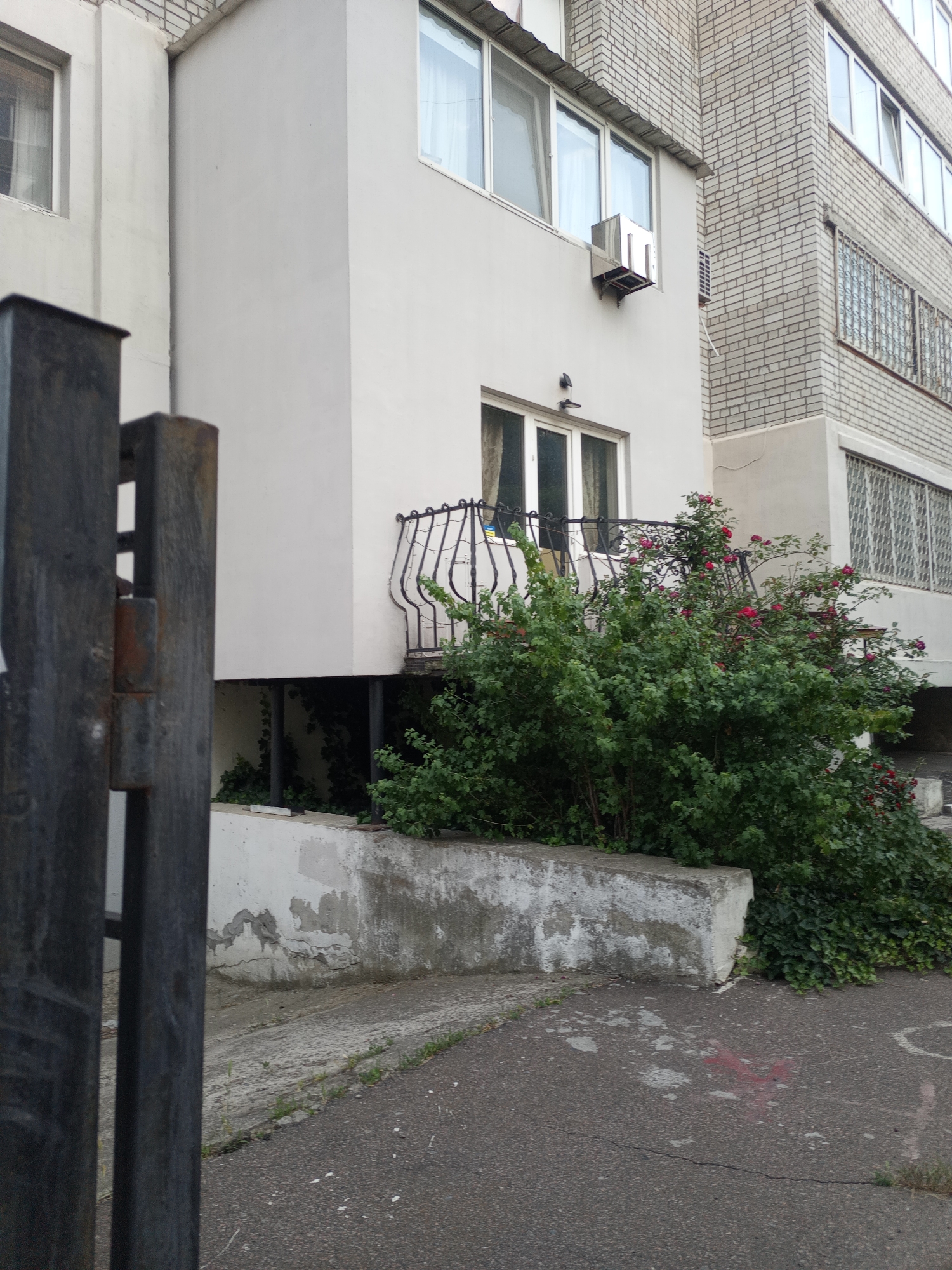 The image shows the exterior of an apartment building in an urban environment. In the foreground, a dark metal gate is partially visible on the left. The building itself is a multi-story structure with a lighter colored facade. There are several windows visible, some with curtains. An air conditioning unit is mounted on the exterior wall. Below a balcony with a decorative railing, a dense green bush with red flowers is growing. The ground in front of the building is paved and shows some signs of wear, with cracks and scattered debris. The surrounding environment appears to be an outdoor urban setting.
