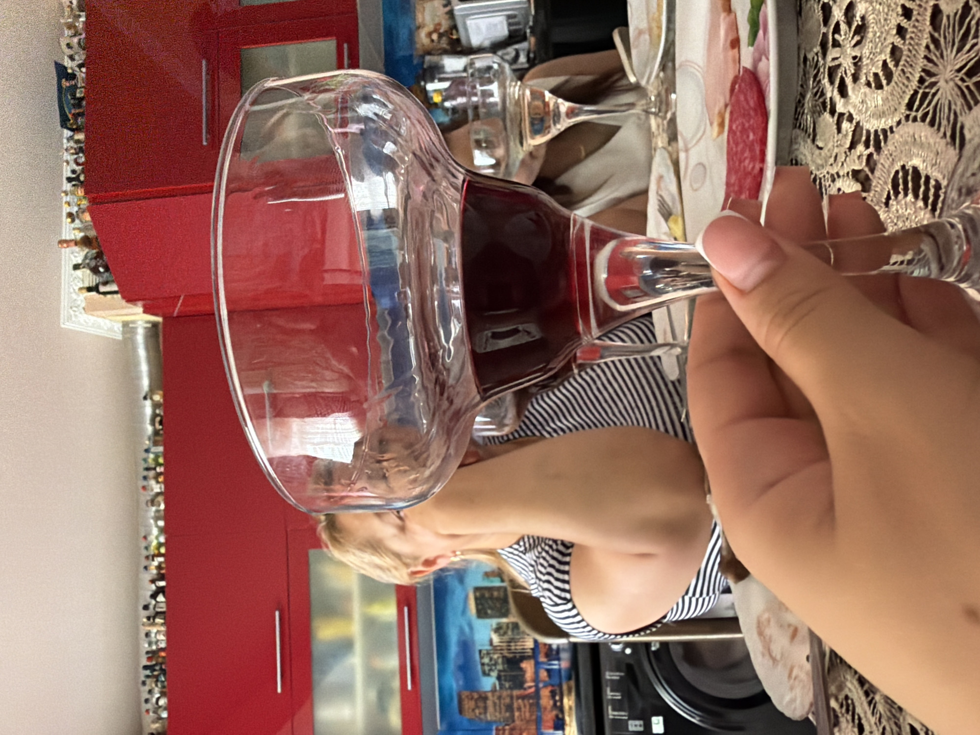 The image depicts a close-up of a hand holding a wine glass filled with red wine. In the background, a woman with blonde hair, wearing a striped shirt, is seated at a table, covering her mouth with her hand. Another person, partially visible, is seated across from her. The setting appears to be a kitchen with red cabinets lining the upper walls, and various bottles are displayed on a shelf above the cabinets. A cityscape mural is visible on the wall behind the seated woman. The table is set with plates of food, including sliced salami and ham. The overall scene suggests a casual meal or gathering in a home environment, potentially located in Durlești, Moldova, given the context provided, though the image itself does not explicitly confirm this location.