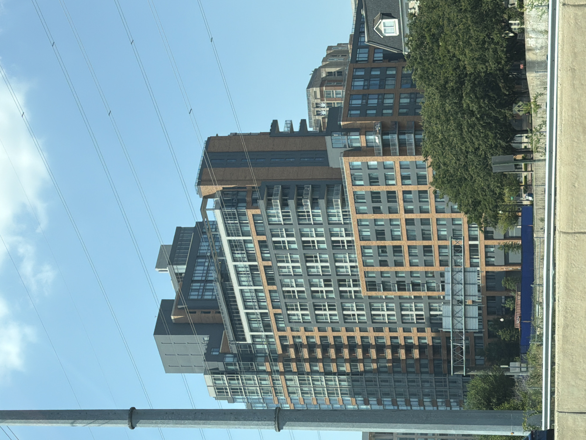 The image shows a tall, modern apartment building in Huntington, United States of America. The building has a complex geometric design with multiple sections of varying heights and colors, featuring a combination of glass windows, gray siding, and orange brick. Several balconies with glass railings are visible, extending from different levels. The building is situated in an urban environment, with a clear blue sky and some white clouds in the background. Several horizontal power lines stretch across the sky in front of the building. To the left, a large, gray utility pole is prominent. In the foreground, a concrete barrier and vegetation are visible, suggesting the vantage point is from a roadway or elevated structure. The overall scene depicts a contemporary urban landscape with a focus on the architectural details of the apartment building.