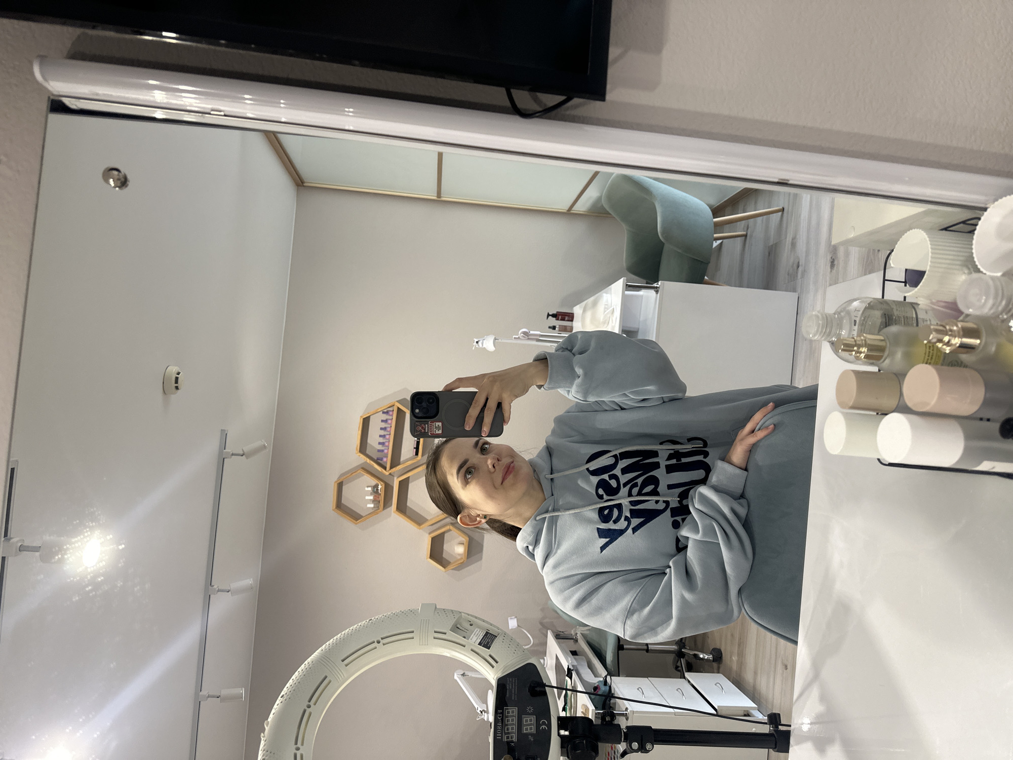 A woman takes a selfie in a room reflected in a mirror. The room contains a ring light on a stand, hexagonal shelves on the wall, a white desk with bottles and containers, and a light green chair in the background. The woman is wearing a light blue hoodie. The photo appears to have been taken in Perm, Russia.