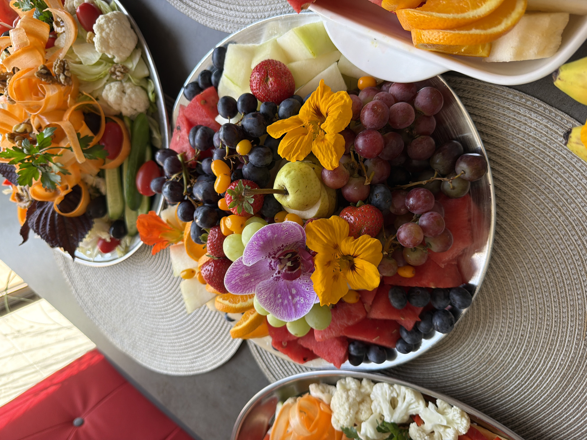 The image shows two fruit platters on a table with a textured placemat. The platter in the foreground is filled with watermelon, grapes (red and green), strawberries, blueberries, orange slices, a pear, and decorated with two orange nasturtium flowers and a purple orchid. The second platter, partially visible in the background, appears to contain a salad with shredded carrots, lettuce, cauliflower, tomatoes, cucumber slices, walnuts, and parsley. To the left of the platters, a red cushioned chair is visible. The scene is indoors, likely in a dining area. There are no people or animals present in the image. The location of the image is not specified in Ventspils, Latvia.