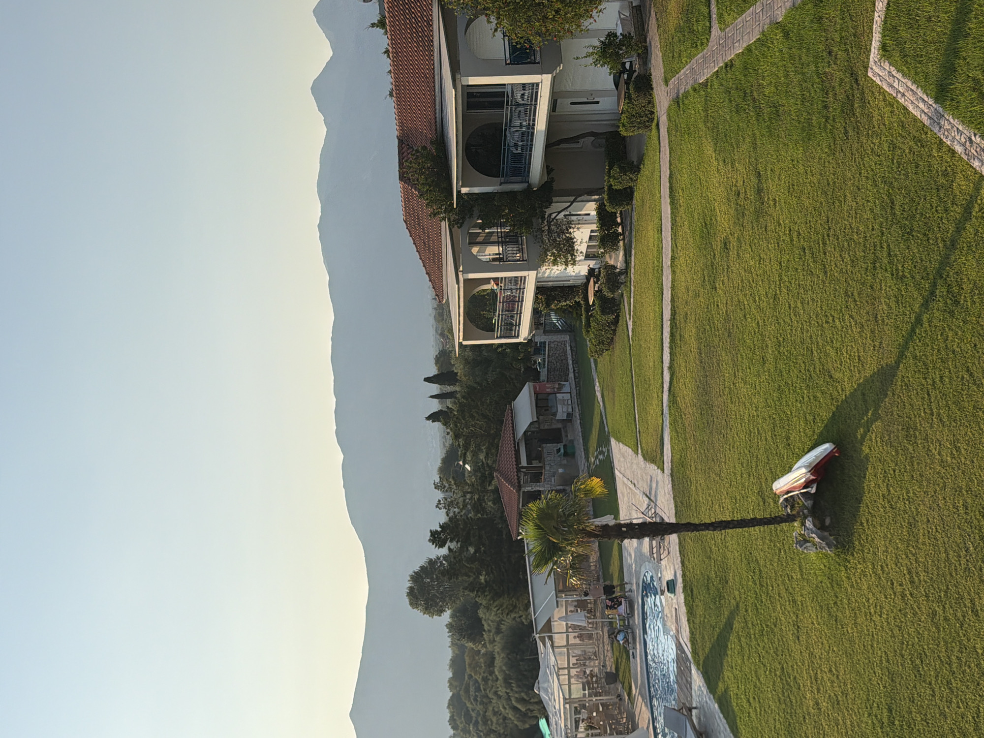 The image depicts a resort area in the Municipal Unit of Alyzia, Greece. In the foreground, a well-maintained green lawn is bordered by paved walkways. A palm tree stands centrally, with two kayaks resting at its base. To the left, a swimming pool with lounge chairs surrounds it, and a covered seating area with tables and chairs is visible with several people seated. Further back, a building with a red-tiled roof and awnings appears to be a bar or restaurant, with patrons and staff present. Adjacent to this, a multi-story building with balconies, white walls, and a red-tiled roof suggests accommodation. In the background, dense green foliage and trees lead up to rolling hills and distant mountains under a clear, pale sky. The overall impression is one of a peaceful, sunlit resort environment.