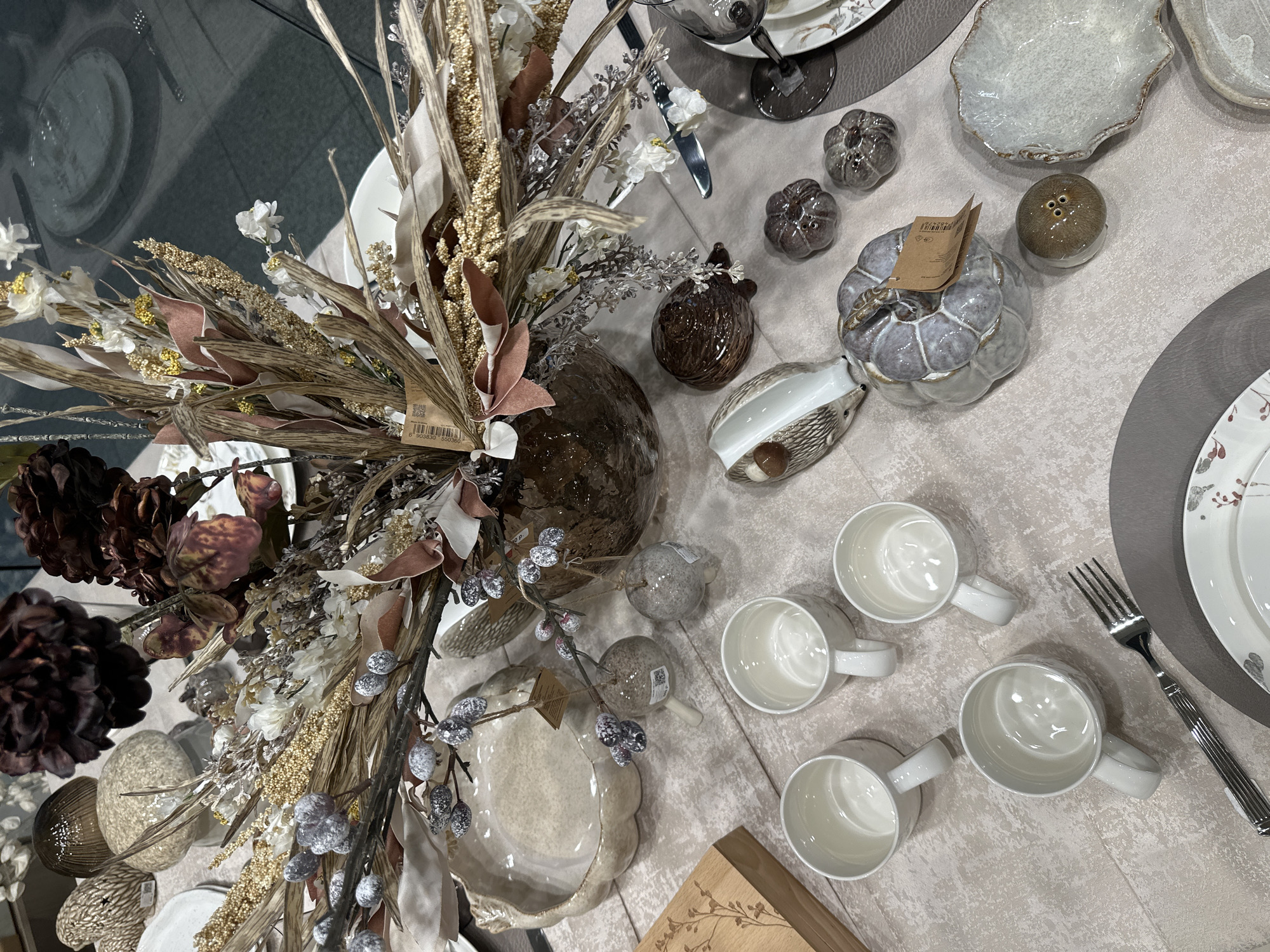 The image depicts a table setting with a decorative centerpiece. The centerpiece is a large arrangement of dried floral elements, branches with faux berries, and some artificial flowers in muted autumnal colors, all contained in a dark, textured vase. Scattered around the centerpiece are various decorative items, including ceramic pumpkins of different sizes and colors, a ceramic hedgehog figurine, and what appear to be small ceramic bells. In the foreground, four white ceramic mugs are arranged, along with a fork. A large, light-colored ceramic bowl with a scalloped edge is also visible, as are other ceramic dishes and placemats. The table itself is covered with a light-colored, textured fabric. The background is blurred, but suggests an indoor setting, possibly a retail display or a home environment. There are no people present in the image. The scene is likely set up for a festive occasion, such as autumn or a holiday gathering. The image does not provide any information about the city of Samara, Russia.