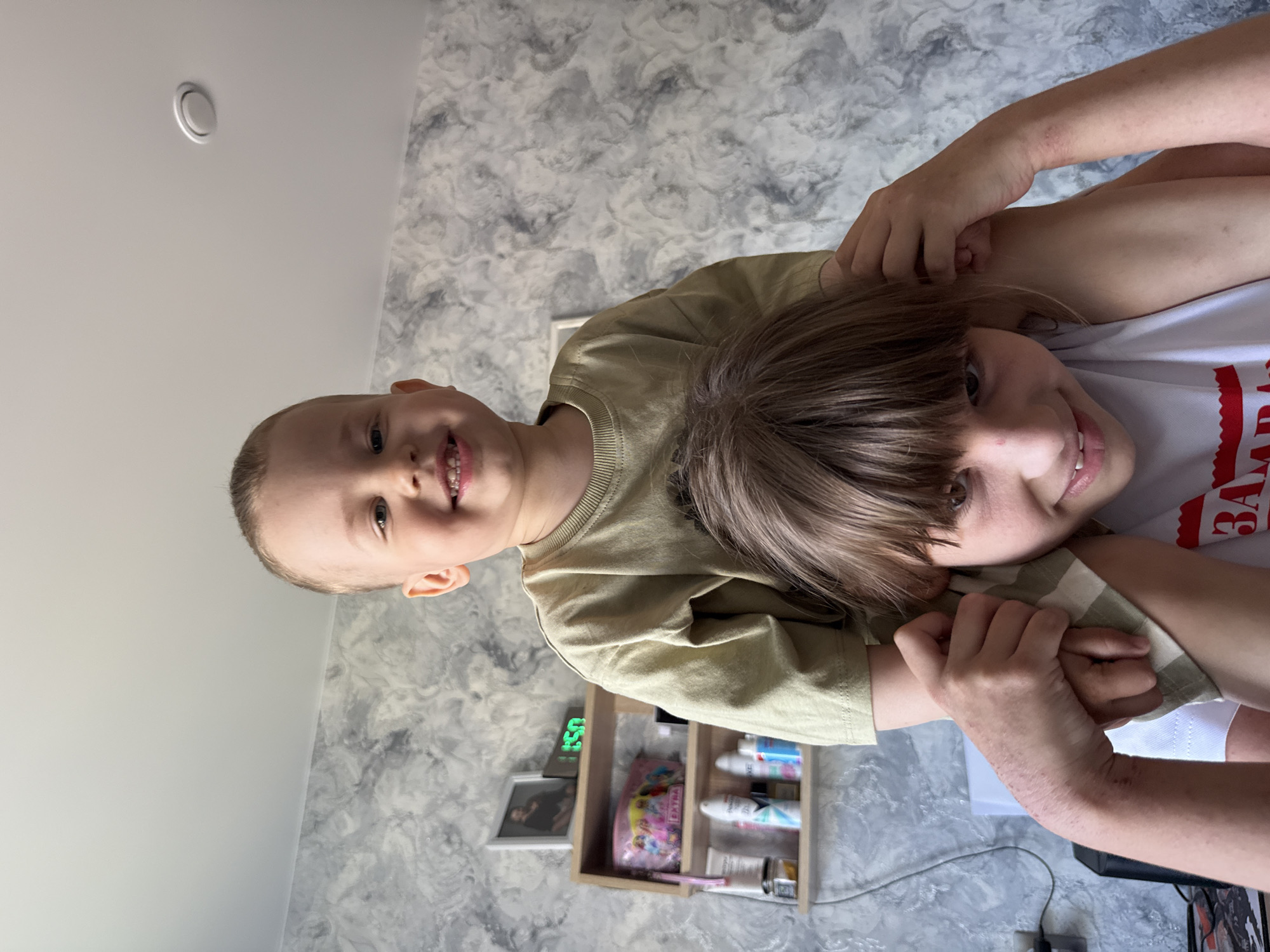 Two children, likely siblings, are posing for a photo indoors. The younger child is sitting on the shoulders of the older child. Both are smiling at the camera. The room has light-colored walls with a patterned wallpaper. A small shelf unit containing various items, including bottles and a pencil case, is visible in the background, along with a digital clock displaying the time '1:50'. The photo was taken at the location of Rusanovo, Russia.