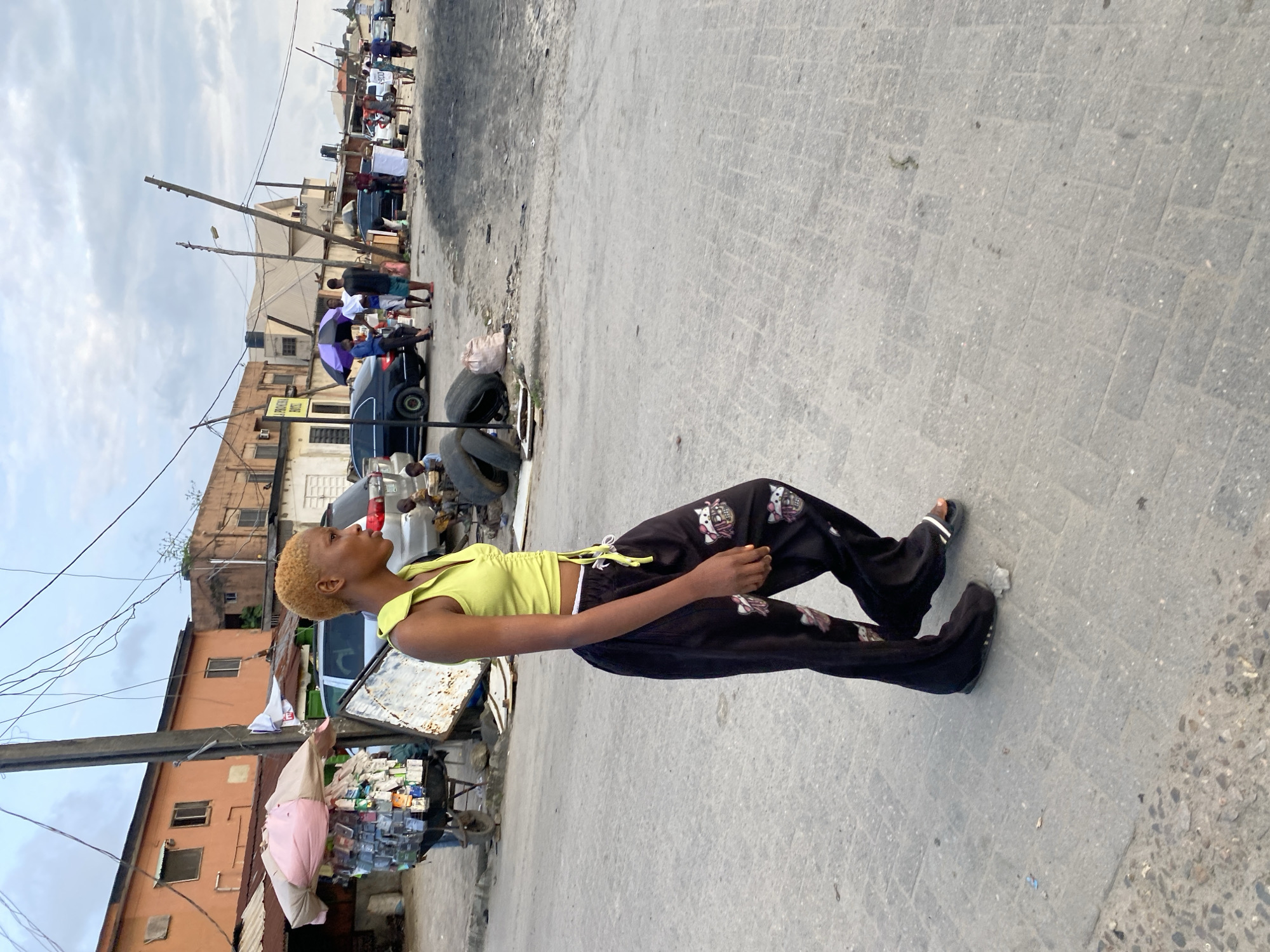 A young woman with short blonde hair walks to the right in the foreground of the image. She wears a sleeveless yellow crop top and loose black pants with a pattern of skulls and flowers. She is on a paved sidewalk. In the background, there are buildings, several parked cars, a cart piled with goods, and other people walking. The sky is cloudy. The scene appears to be an outdoor market or street in a city.