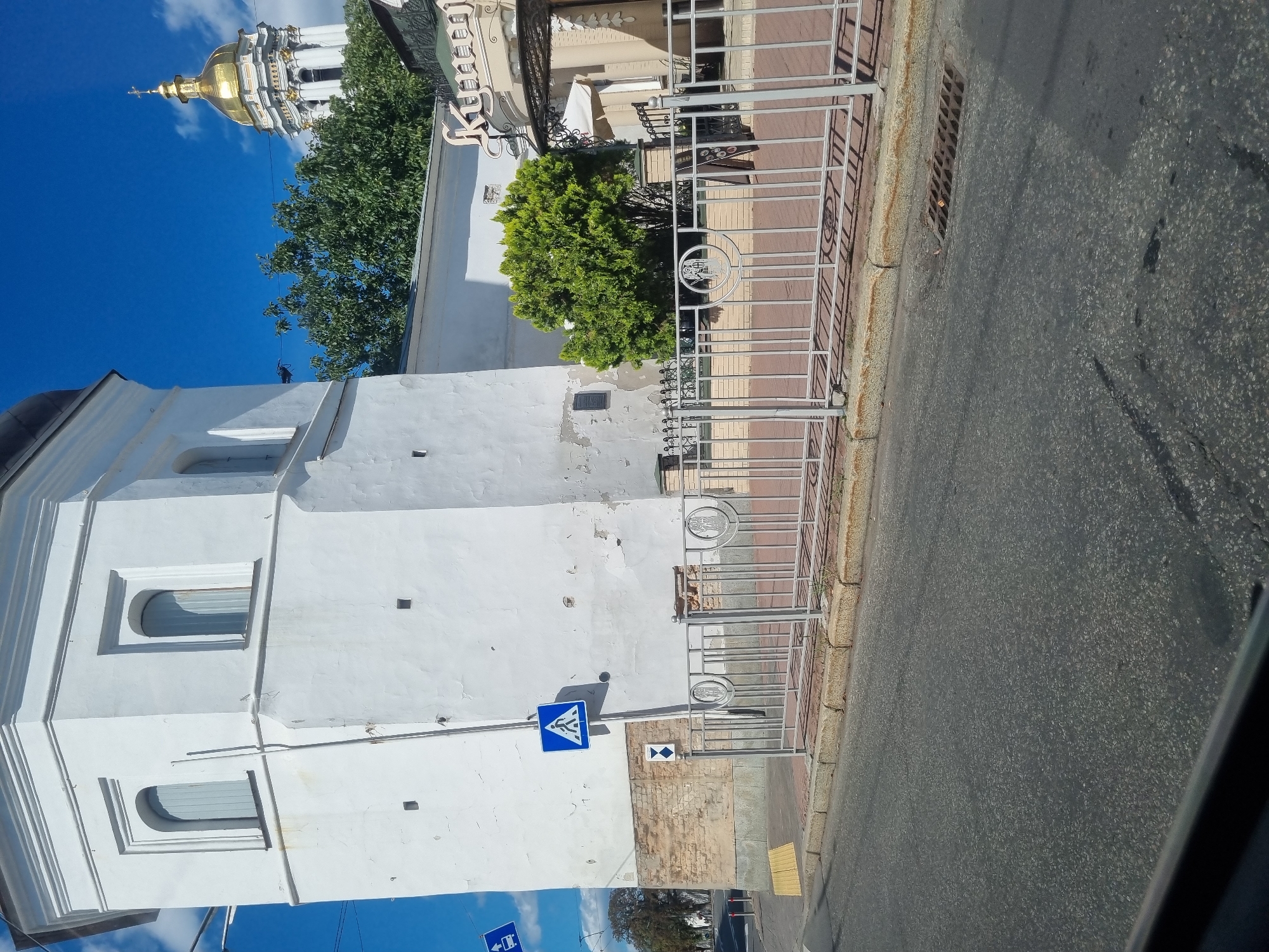 The image shows a view of the Lavra Bell Tower and the Church of the All Saints in Kyiv, Ukraine, on a clear, sunny day. In the foreground, a street is visible with a grey asphalt surface and a sidewalk with a yellow tactile paving. A white metal fence with decorative circular elements lines the sidewalk. A pedestrian crossing sign is affixed to the side of a tall, white, octagonal tower. Behind the fence, a small, lush green tree stands in front of a building with a large, ornate sign that appears to read "Кулинар". In the background, the golden dome of the Church of the All Saints gleams against the bright blue sky. The overall scene suggests a vibrant urban environment with historical and religious landmarks.
