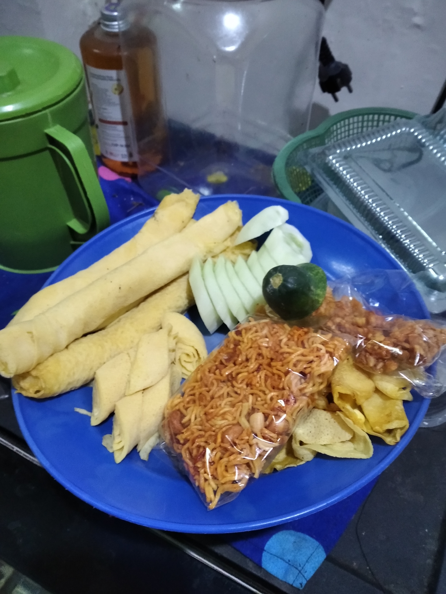 A blue plate of Indonesian food is on a dark surface. The plate contains several pieces of what appear to be fish cakes, sliced cucumber, a lime, and two bags of fried snacks. One bag contains what looks like fried noodles, and the other bag contains chips. In the background, there is a green pitcher with a handle, a clear container, and a green basket containing a silver tray. The lighting suggests an indoor setting, possibly a home or a small eatery in Rambipuji, Indonesia. There are no people visible in the image.