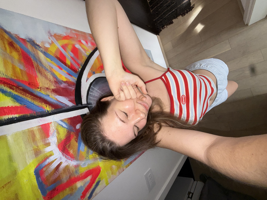 An indoor scene in Vozdovac Urban Municipality, Serbia, depicts a young adult female from a high-angle perspective. The female has light skin, long brown hair, and appears to be smiling with eyes directed downward. She is wearing a red, white, and light blue horizontally striped spaghetti strap crop top with a white oval patch on the chest displaying the word "NINETIES" in red text. She also wears light blue and white striped shorts. Her left arm is extended towards the camera, indicating a self-portrait. Her right hand is formed into a fist, resting near her right cheek. The background features a large, colorful abstract painting on a light-colored wall, primarily composed of red, yellow, blue, black, and white brushstrokes, with a prominent black and white circular element on the right. The floor is light-toned wood or laminate. A dark, textured rug is partially visible on the right, near what appears to be a dark doorway or furniture. A white electrical outlet or light switch is present on the left wall. The area is well-lit.