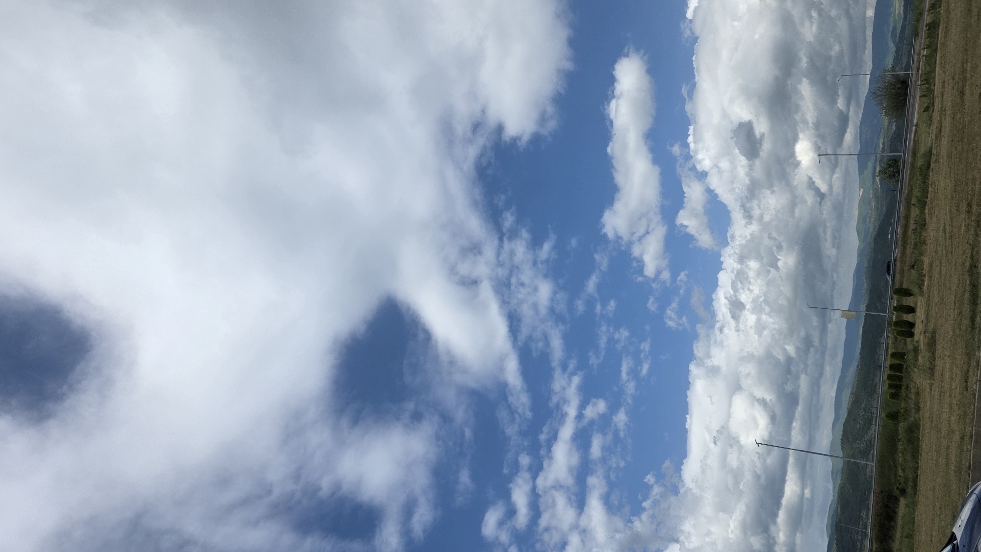 The image depicts a landscape scene under a sky with a mixture of cloudy and clear blue sections. The right side of the frame shows a grassy field with some vegetation, possibly trees or bushes, along a line that could be a road or border. The landscape appears hilly or mountainous in the background. In the sky, there are clouds varying in density, from thin, wispy formations to thick, cumulonimbus clouds. There are also lampposts. The image is taken outdoors, with natural lighting. The location is potentially near Blagoevgrad, Bulgaria, as requested.