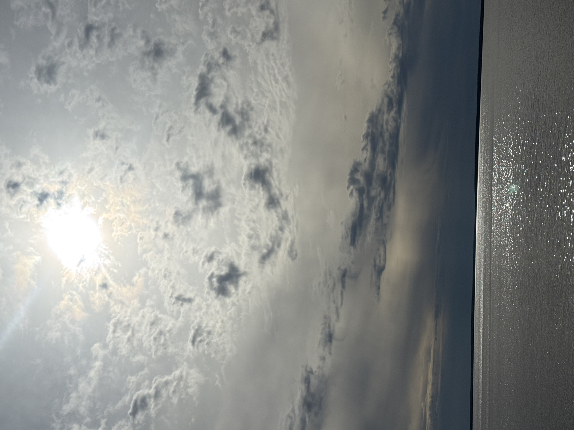 The image shows a view of the sky over water. The sky is mostly covered in clouds, with the sun shining through in the lower left corner. The water on the right is reflective and appears to be shimmering. The location is Ivanovo, Russia near the Timiryazevskii S/S.