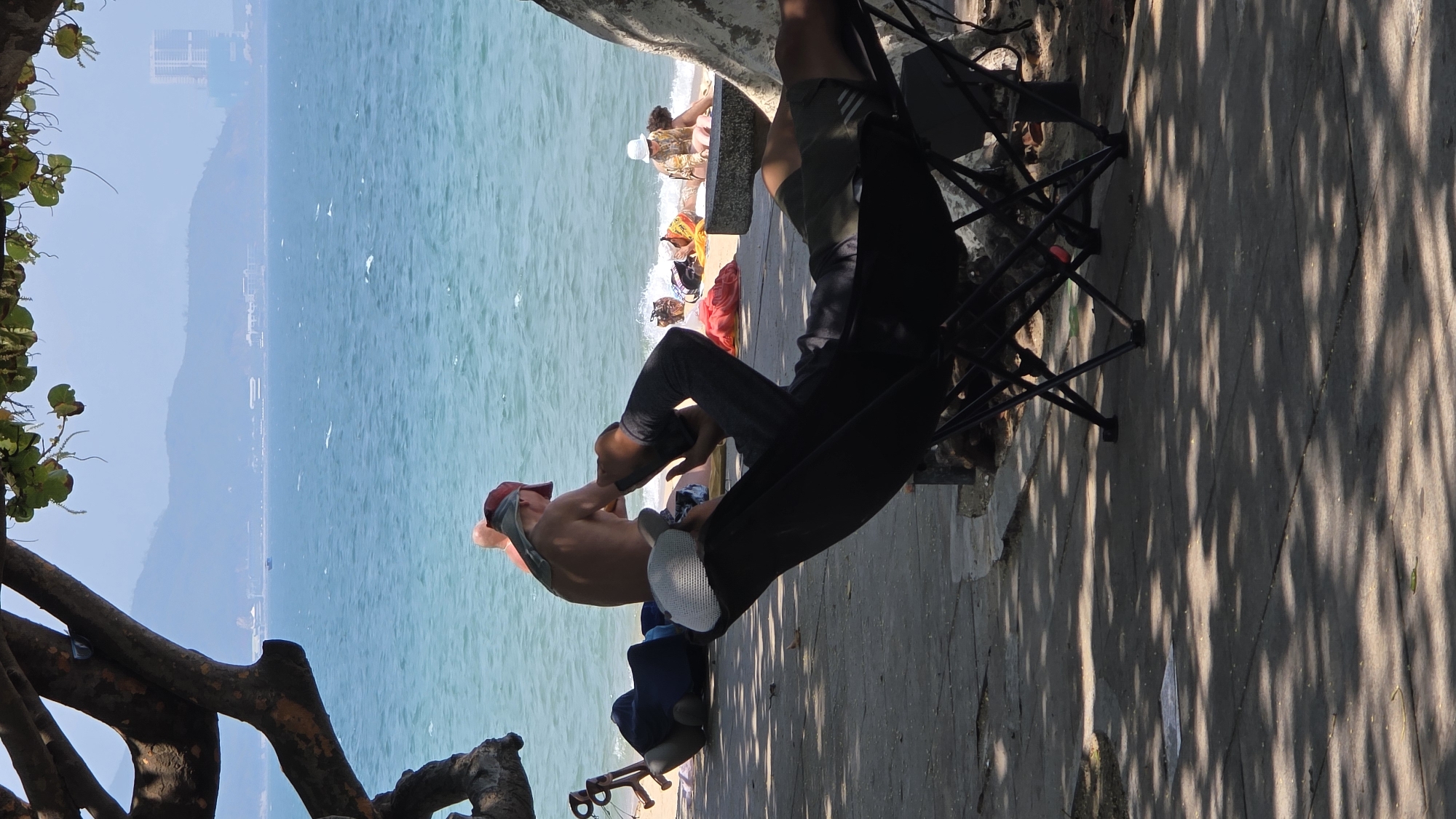 The image depicts a scene at a beach in Nha Trang, Vietnam. The water appears blue, and the shoreline is visible with people relaxing on the sand. A man wearing a red cap and no shirt is seen sitting by the shore, apparently talking to another man in a black shirt and pants seated on a portable chair. The sun is shining, creating shadows on the paved area next to the beach where the men are situated. A tree with exposed branches is visible on the side, partially obscuring the view. People are gathered on the shore beyond. A building, likely a hotel, is visible in the background.
