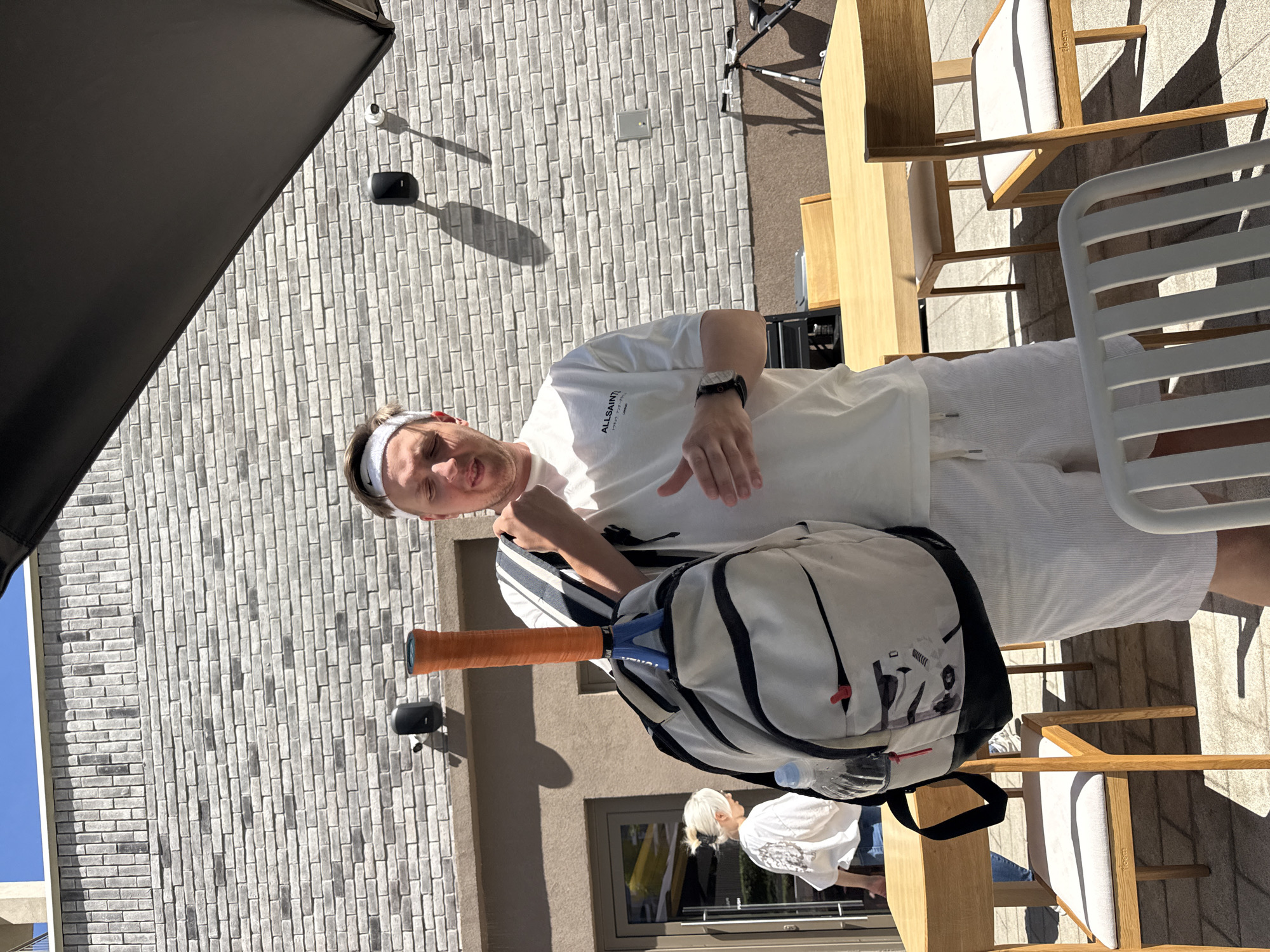 A man stands outside on a patio, holding a tennis racket and bag. He wears a white t-shirt with "ALLSAINTS" printed on it, a white headband, white shorts, and a watch. Behind him is a light gray brick wall with two dark gray security cameras attached. Nearby are wooden patio chairs and a wooden table. A woman with blond hair pulled back is walking through a doorway behind the man. It is sunny. The location is likely in Belgrade, Serbia.