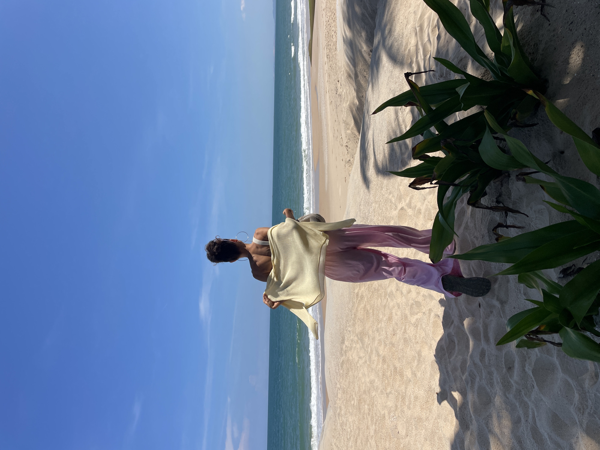 A person stands on a sandy beach, facing the ocean. They are wearing pink wide-leg pants and a light yellow cardigan draped over their shoulders. The person's back is to the camera, and their hair is pulled up. The ocean is calm with small waves. The sky is clear and blue. In the foreground, there are shadows cast by palm trees on the sand and some green plants. The location is identified as Phu Thuy Ward, Vietnam.