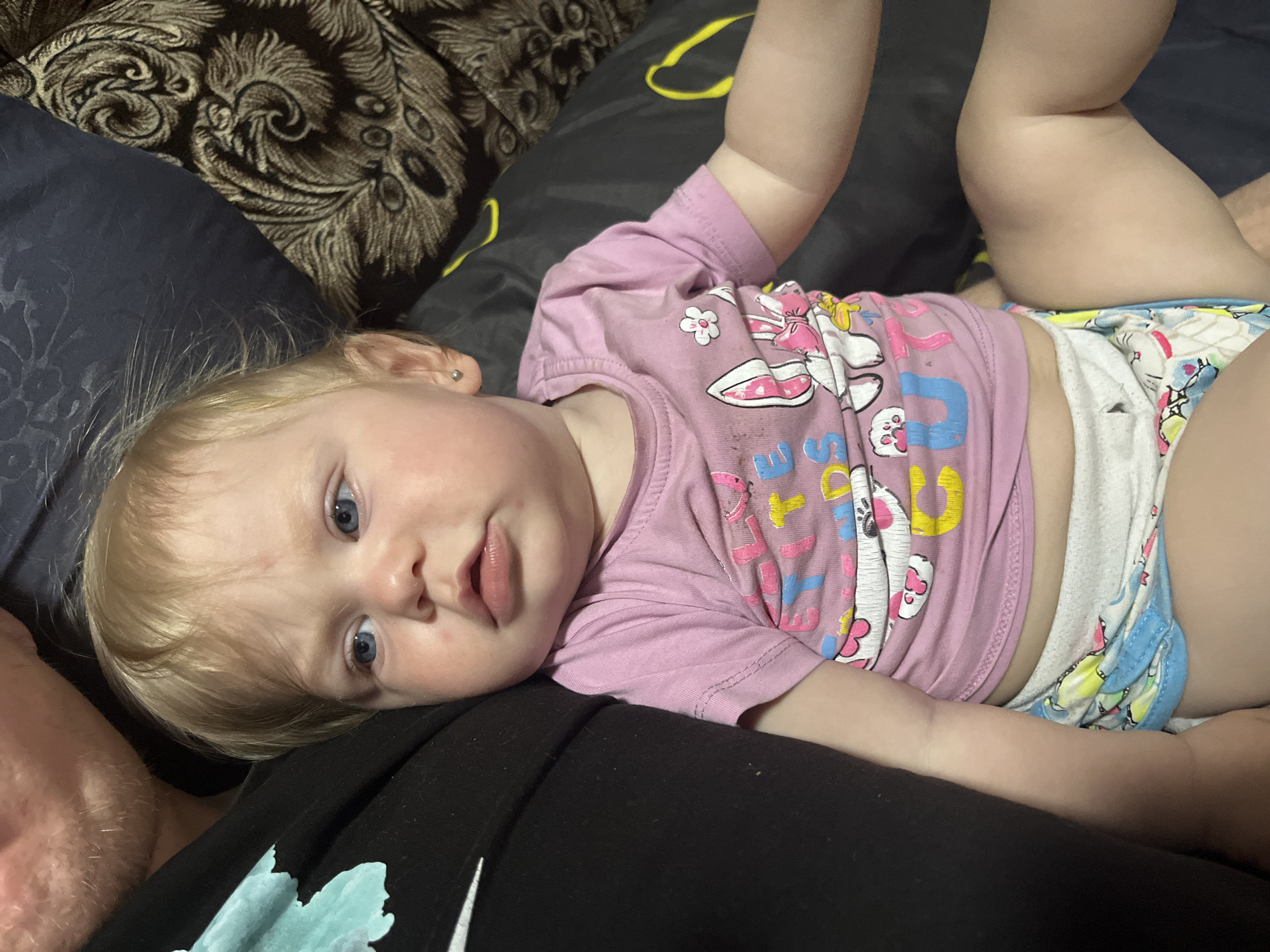 A close-up, high-angle shot captures a young child lying on their back, propped up by dark fabric. The child has fair skin, blonde hair, and blue eyes, with a slightly open mouth. They are wearing a pink t-shirt with cartoon graphics, including what appear to be rabbits and flowers, and a pair of patterned underwear. The background consists of dark cushions and a dark patterned fabric. One cushion has a light-colored, ornate pattern. The setting appears to be indoors, possibly a home, given the presence of soft furnishings. The child's expression is neutral, with their gaze directed towards the camera. The lighting suggests it is daytime, but there are no strong indicators of the time of day or weather conditions. The location context provided is Фоки, Russia. There is no visible text in the image.