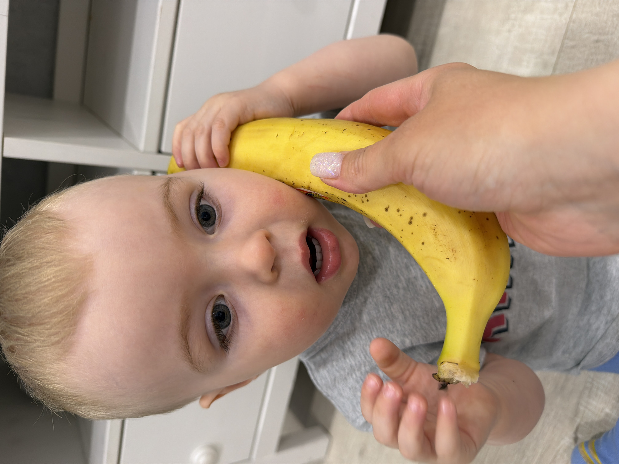 A toddler with blond hair lies on their back next to a white cabinet. An adult hand holds a banana against the toddler's face as if it's a telephone. The toddler looks at the camera with their mouth open. The location is indoors. The city of Votkinsk, Russia, cannot be verified from the image.