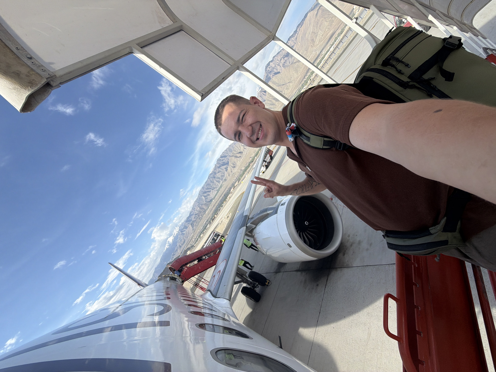 A young man with short hair and a brown shirt takes a selfie as he stands on the tarmac next to an airplane, mountains in the background. The man is wearing a backpack and makes a peace sign with his hand. He is partially obscured by a covered walkway. A red piece of equipment is in the foreground. An airplane with red accents is to the left of the man. The sky is blue with scattered clouds. The man's location is the city of Leh, India.