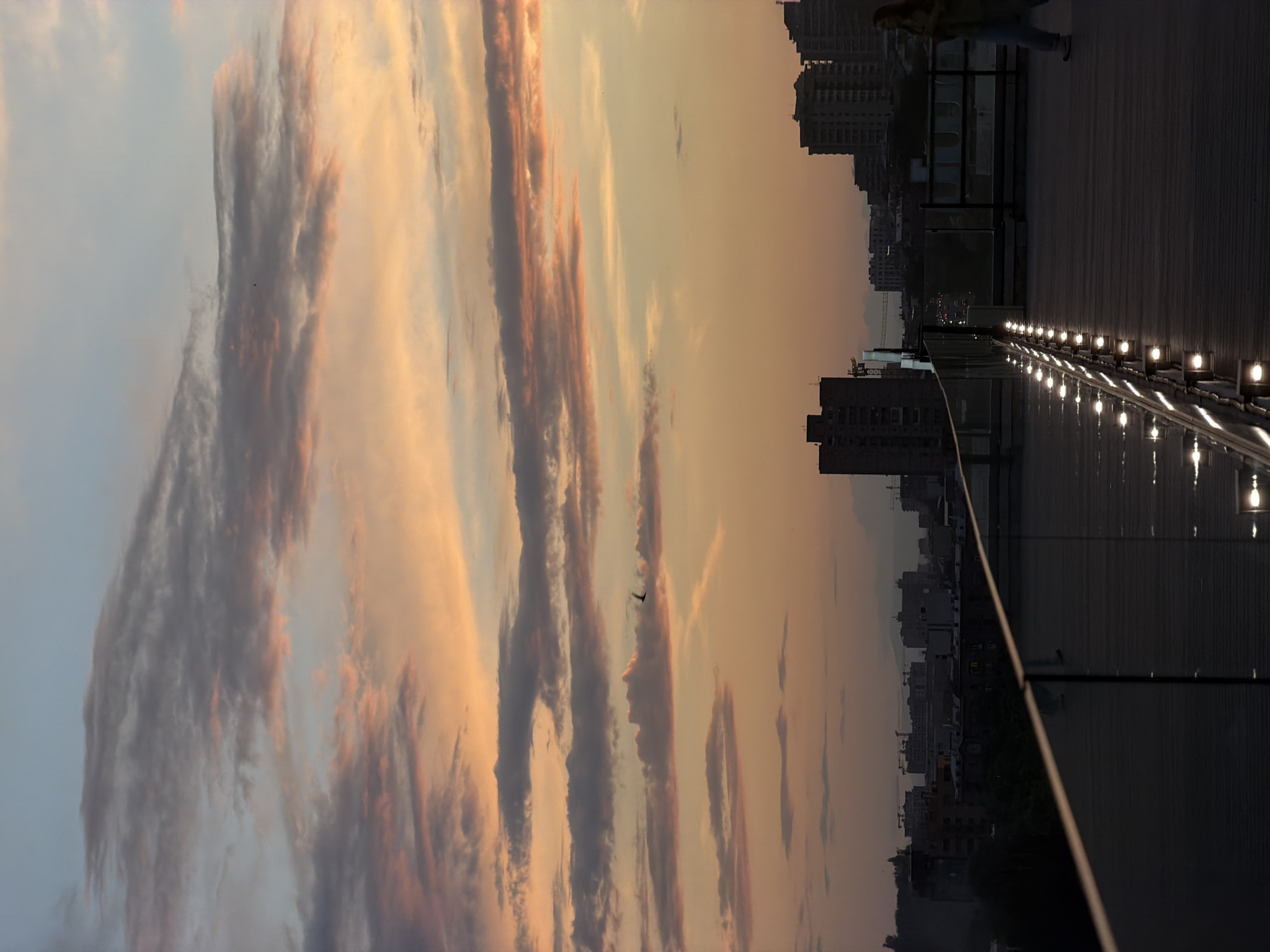 The image captures a sunset scene over the city of Yerevan, Armenia, observed from an elevated perspective, likely a rooftop or balcony. The upper two-thirds of the frame are dominated by the sky, which transitions from a pale blue at the zenith to warm orange and pink hues near the horizon, indicative of late dusk. Overcast conditions are present, with multiple layers of grey, pink, and orange clouds horizontally stratified across the sky. Two small, dark birds are visible in flight within the sky portion.
The middle ground features the silhouette of the cityscape, consisting of numerous multi-story buildings and visible construction cranes, suggesting urban development. Faint outlines of mountains are discernible in the distant background.
The foreground, occupying the lower third of the image, depicts a dark, flat rooftop surface. A modern railing, constructed with glass panels and a dark top rail, extends diagonally from the lower left towards the mid-right. Along the base of this railing, a series of evenly spaced, circular white lights emit distinct points of illumination, casting reflections on the railing and the dark floor.
A single individual is present on the rooftop, walking towards the right side of the frame. The person is partially silhouetted and viewed in profile, wearing light-colored trousers and a dark top or jacket. Their facial features and specific attire details are indistinct due to the low light.