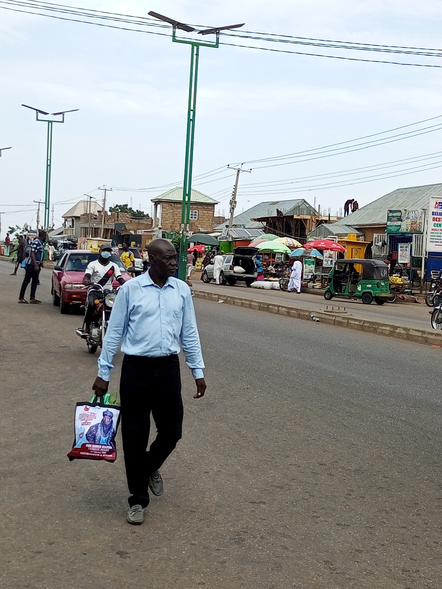 The image depicts a street scene in Gombe, Nigeria. In the foreground, a man wearing a light blue collared shirt and dark trousers walks towards the right, carrying a patterned shopping bag. Behind him, a motorcycle carries a rider wearing a white t-shirt with black trim and a face mask, and another person wearing a dark t-shirt. A red car is stopped on the left side of the road. The street is paved and lined with various buildings, some appearing to be shops or market stalls, with green and red umbrellas providing shade. Several other individuals and vehicles are visible in the background, suggesting a busy urban environment. Overhead, power lines and tall street lights with solar panels are present. The sky is overcast.