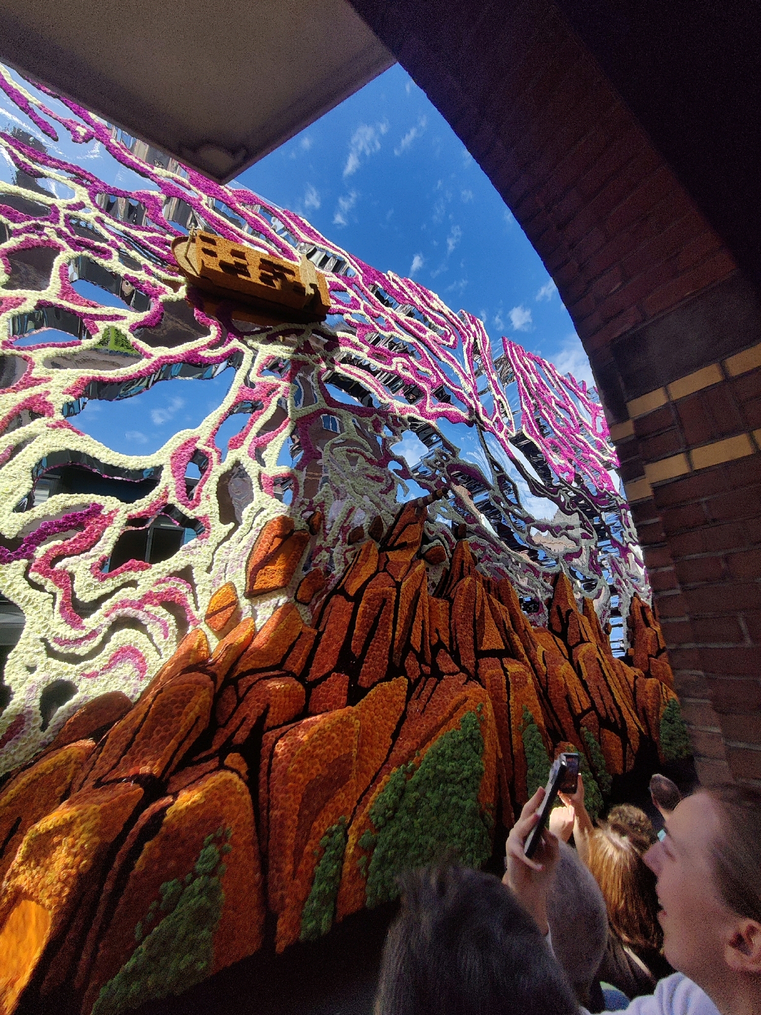 The image depicts a colorful and elaborate float, likely part of the Zundert Flower Parade, situated within the city of Zundert, Netherlands. The float is composed of a mountainous landscape rendered in shades of orange and brown, adorned with textured floral elements. Above the mountainous base, a swirling, abstract structure of pink and cream-colored floral elements is attached to a highly reflective surface that mirrors the blue sky and white clouds. A small wooden boat is positioned on this reflective structure. In the foreground, several individuals are visible, with one person holding up a smartphone to capture a photograph or video of the float. The scene appears to be viewed from below, with the archway of a building framing the top of the image. The architecture suggests an urban or semi-urban setting.