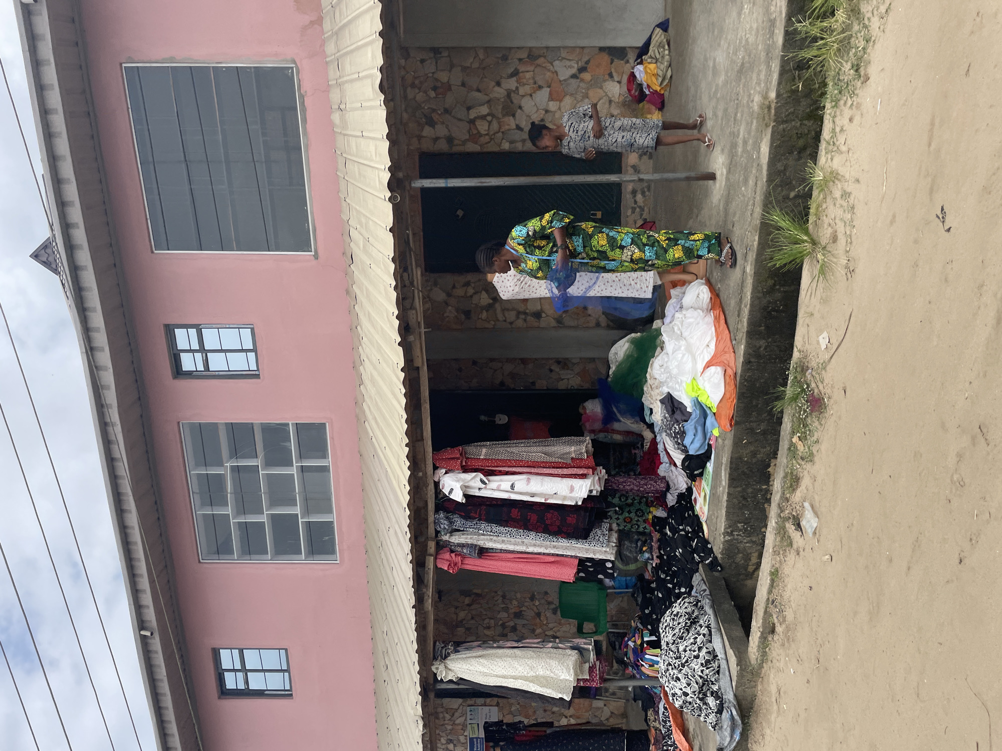 In Badagry, Nigeria, a pink building with multiple windows, some with divided panes and others with dark, slatted coverings, stands above a covered outdoor market stall. The stall's roof is corrugated metal, supported by wooden beams. Inside the stall and spilling onto the concrete walkway in front are piles of clothing and fabric, including dresses on hangers and stacks of patterned textiles. A green plastic chair is visible. Two women are present: one in a vibrant, patterned green and yellow dress, standing and holding what appears to be measuring tape or a string of beads, and a younger girl in a patterned gray dress and flip-flops, standing to her right. The ground in front of the stall is a sandy dirt road. Overhead, power lines stretch across a cloudy sky.