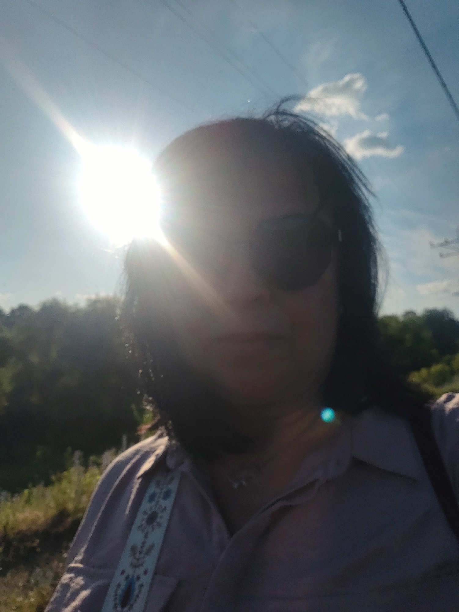 A dimly lit, selfie-style photograph captures a person from the chest up. The individual, facing forward, wears sunglasses and a collared shirt with an embroidered strap. The sun, positioned in the upper left quadrant of the image, is the primary light source, causing lens flare and obscuring facial features. The background consists of trees and a bright blue sky with scattered clouds. The location is identified as Unknown, Unknown.