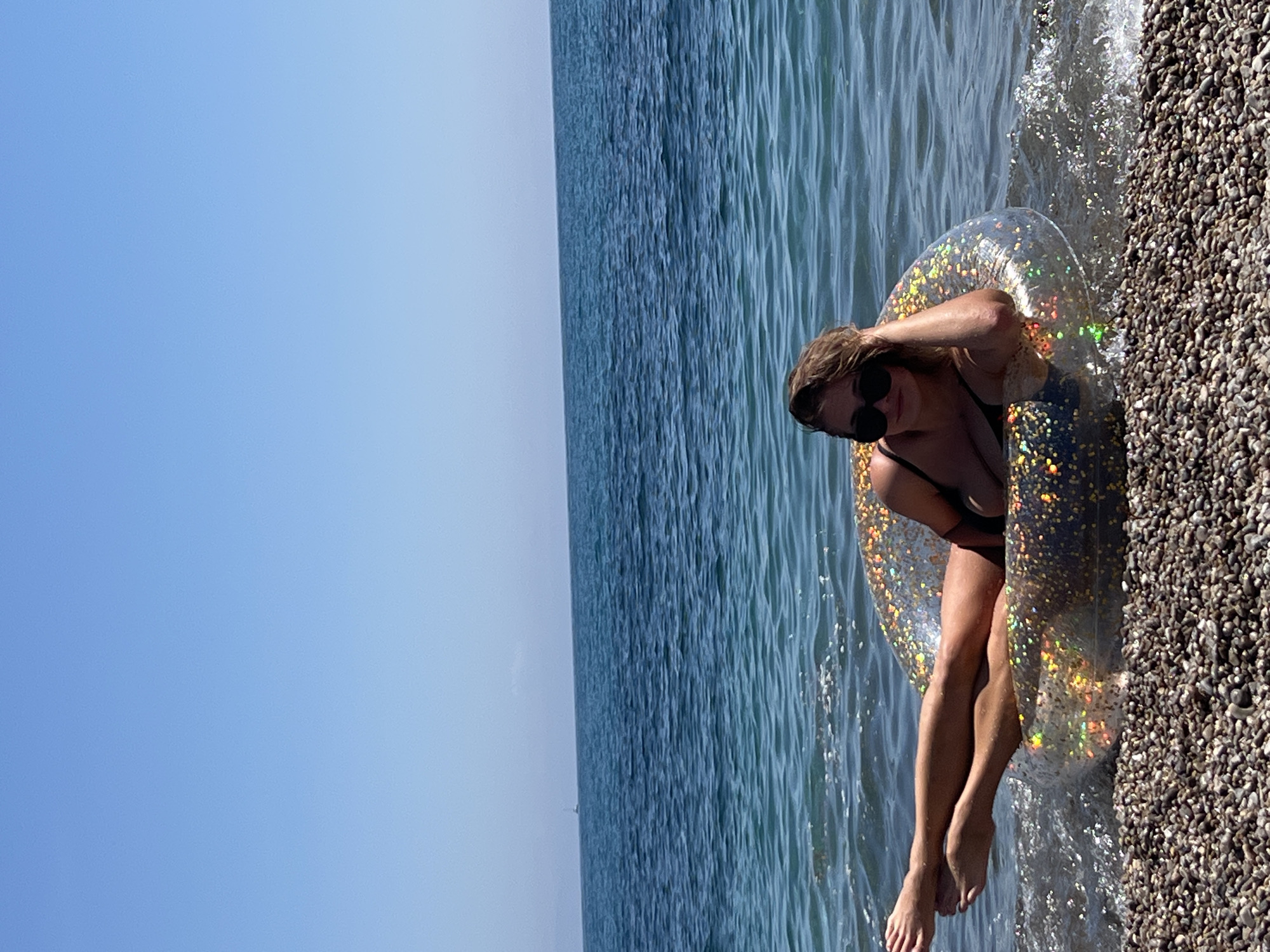 The image captures a woman relaxing in the clear, blue-green sea at a pebbled beach in Prijevor, Montenegro, under a bright, clear sky. The woman, with light brown hair and wearing sunglasses and a dark bikini top, is reclining in a transparent inflatable ring. The ring is filled with glitter or holographic specks that catch the sunlight, creating vibrant, multicolored reflections. Her legs are extended into the shallow water, which gently ripples against the dark grey pebbles of the shore. The expansive, calm sea stretches out to the horizon, where it meets a pale blue sky. A small, faint object, possibly a distant boat, can be seen on the horizon. The bright sunlight indicates it is daytime, likely midday or early afternoon, conveying a sense of leisure and enjoyment during warm weather. There is no visible text in the image.