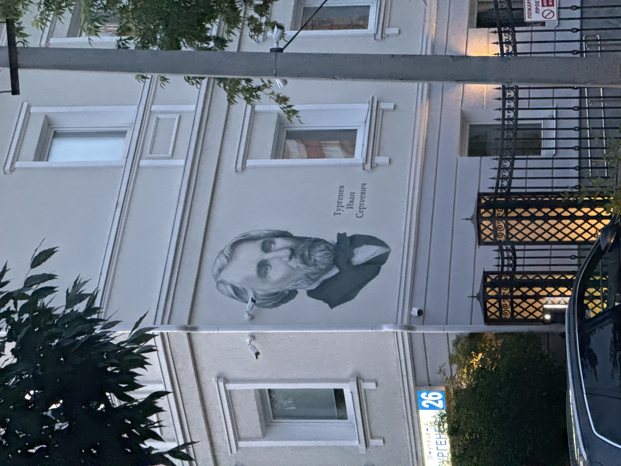 The image depicts a mural of Ivan Sergeyevich Turgenev on the side of a building, located at Turgenev Street, building 26, in Yekaterinburg, Russia. The mural is a black and white portrait of Turgenev, a renowned Russian writer, positioned on the building's facade. The building appears to be a residential or commercial structure, with several windows visible. Security cameras are mounted on the exterior of the building near the mural. In the foreground, a black car is partially visible, and a decorative metal fence with illuminated posts stands in front of the building. The environment suggests an urban setting, possibly during the evening or dusk, given the lighting conditions and the illuminated fence.