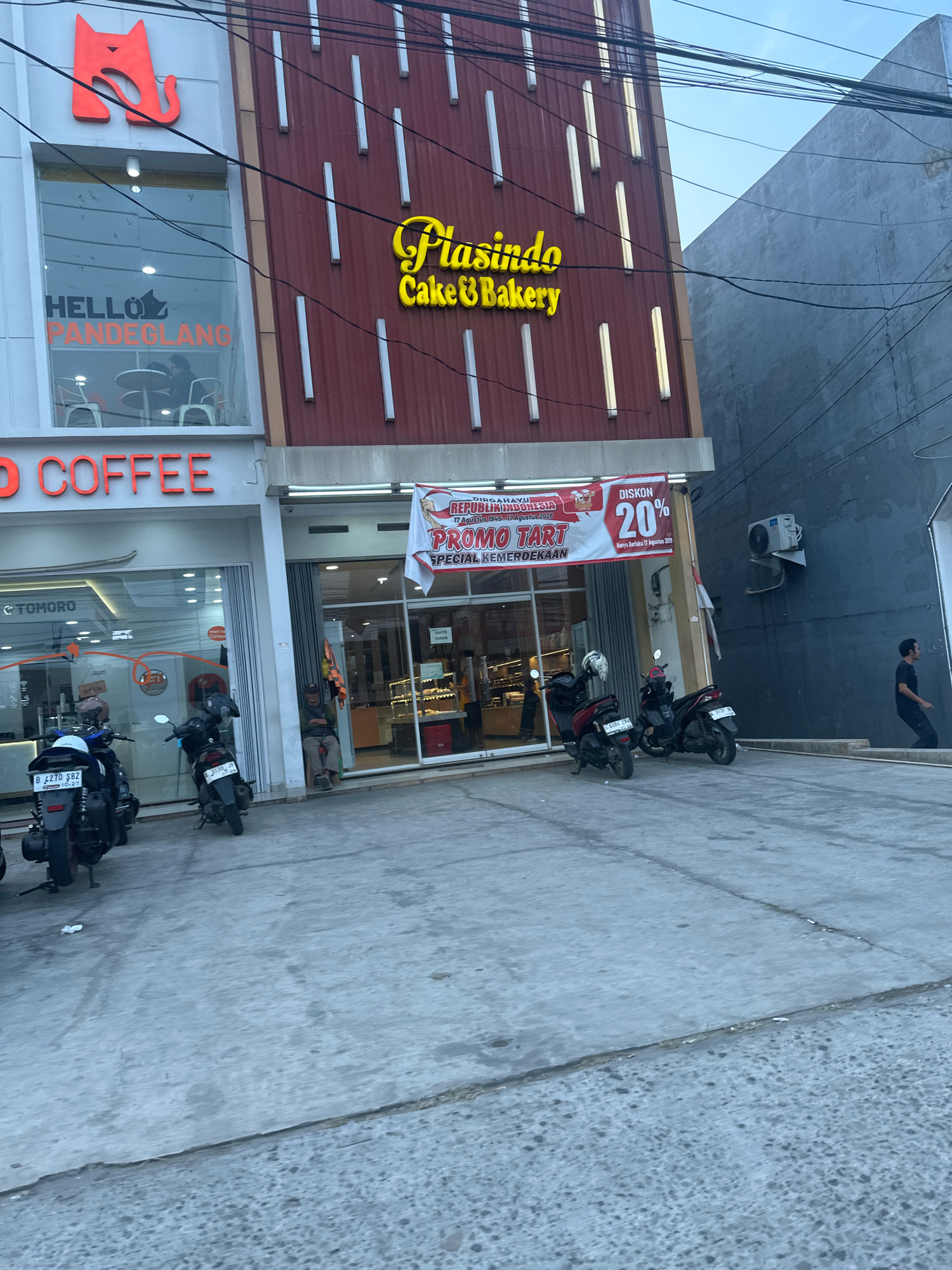 The image depicts the exterior of a commercial establishment, likely a bakery and coffee shop, located in Saruni, Indonesia. The facade features a prominent red cat logo on the left, with the words "HELLO PANDEGLANG" visible through a window, suggesting a connection to the Pandeglang region. Next to it, a red banner announces a "PROMO TART" for "DIRGAHAYU REPUBLIK INDONESIA" (Indonesian Independence Day), with a "DISKON 20%" (20% discount). The building's facade is primarily a dark red metal with vertical white LED lighting.
In front of the establishment, several motorcycles are parked on a concrete pavement. A person is seated on a chair outside the shop entrance on the right side. Further to the right, a dark-colored building with a visible air conditioning unit stands adjacent. Another individual in a dark shirt is walking away from the building on the right. The scene is outdoors, under a bright sky with visible electrical wires overhead.