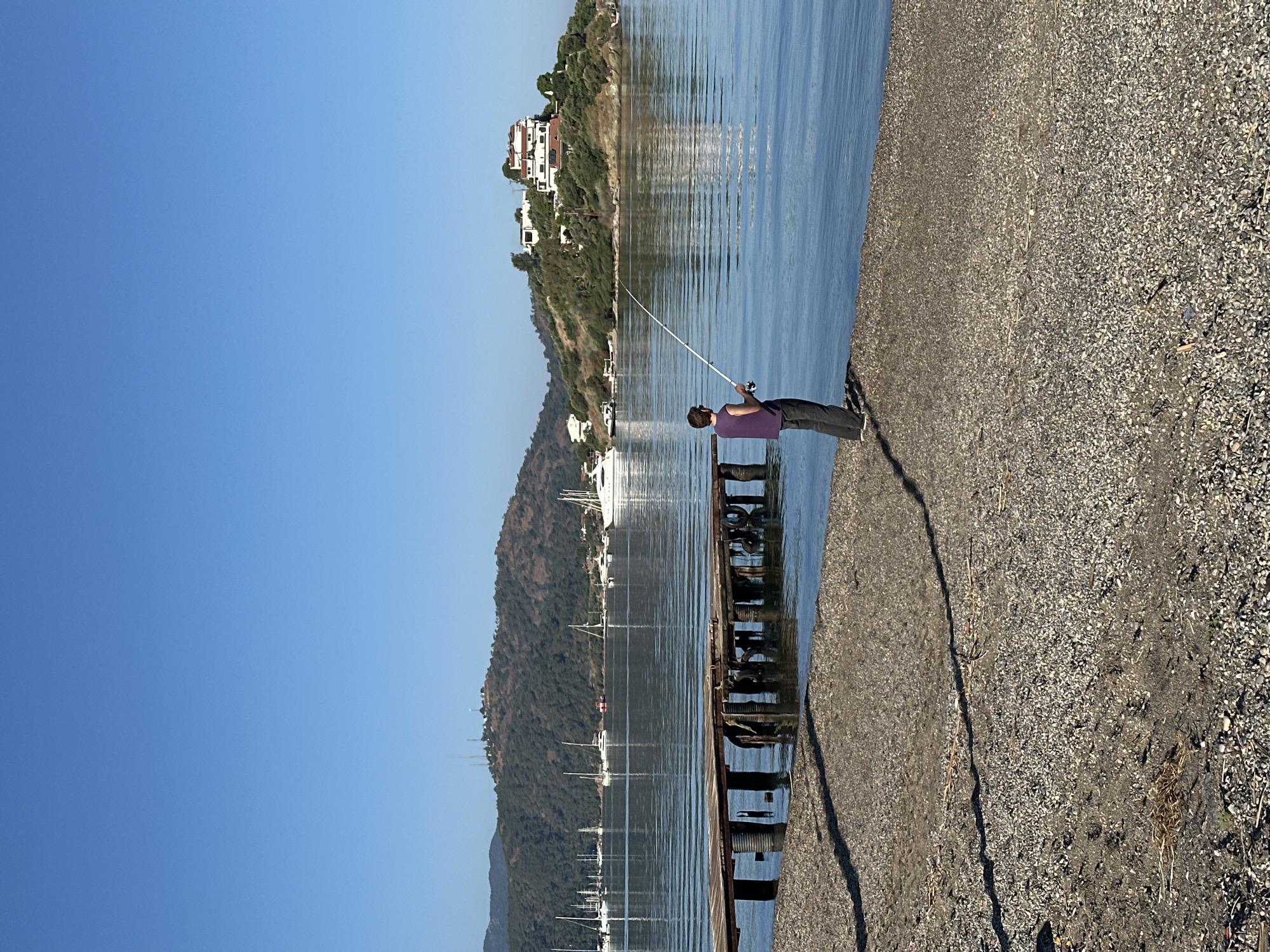 A man in a purple shirt fishes on a stone beach near Fethiye, Turkey. The location features clear blue sky, mountains, and a calm body of water. Boats are seen on the water. A building sits at the top of a cliff. A pier is in the water near the fisherman.