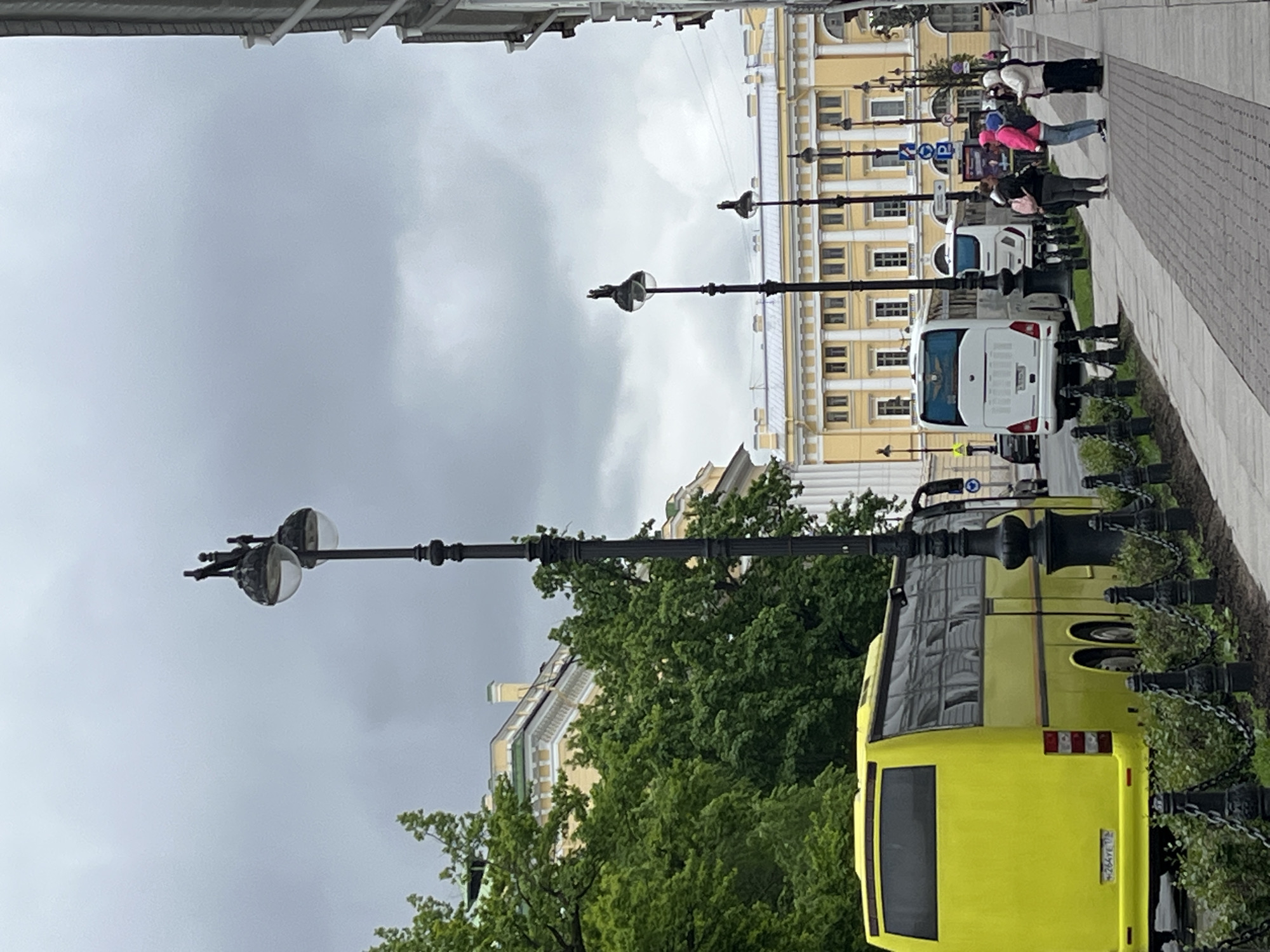 The image shows a street scene in Saint Petersburg, Russia. A yellow bus is parked on the right side of the street. White vans are also parked further down the street. Pedestrians are walking on the sidewalk. Ornate street lamps line the street. Buildings with light yellow facades are visible in the background. The sky is overcast. A metal overhang is visible at the top of the frame. A decorative metal fence is also on the right side of the frame. The street is paved.