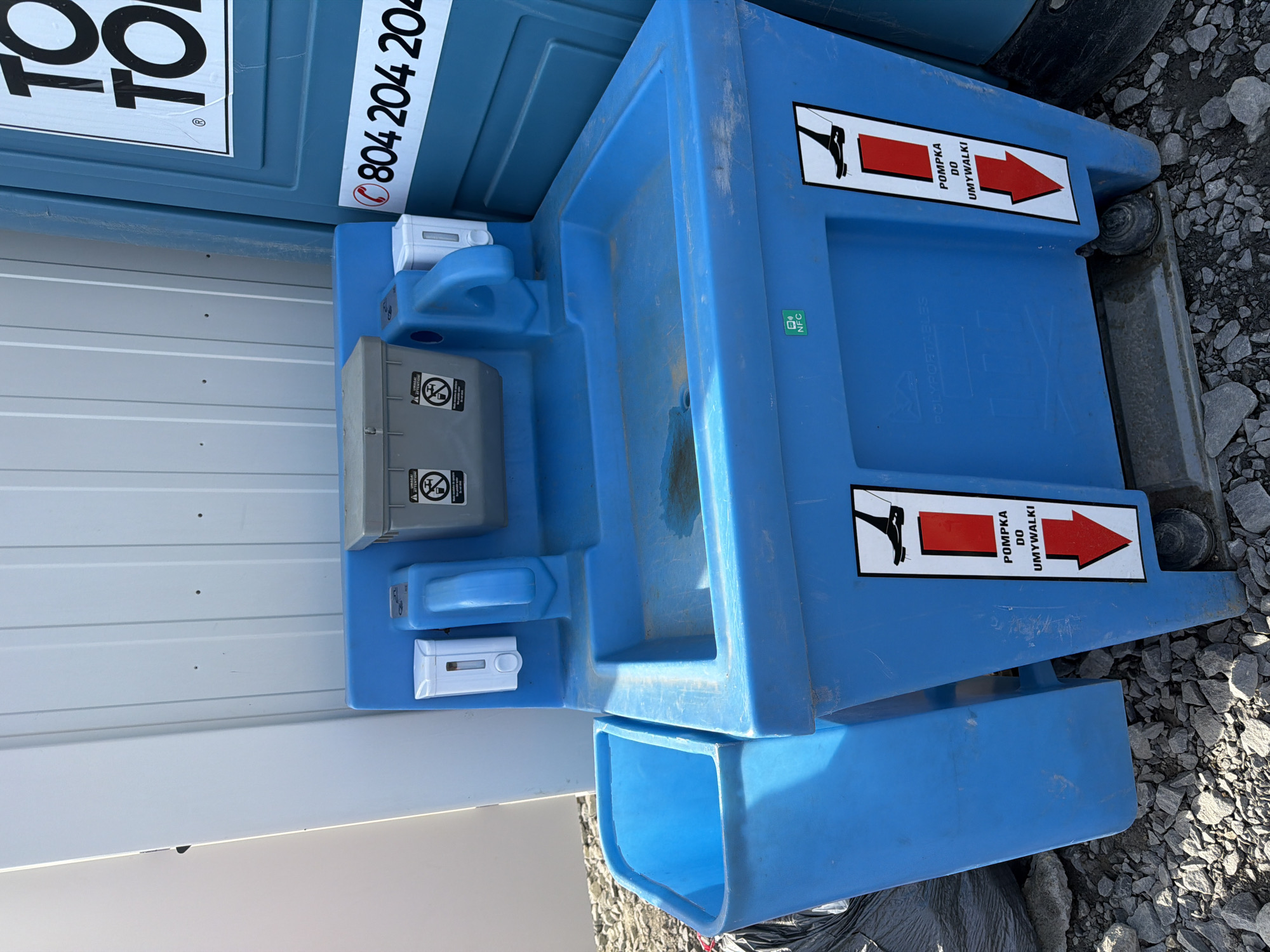 The image shows a blue portable sink unit situated outdoors on a surface covered with gravel. The sink unit is made of plastic and features a basin, two soap dispensers mounted on either side of a central handle, and a grey covered container on top. On the right side of the sink unit, a blue portable toilet is partially visible. A white sticker with a red phone symbol and the text "0804 204 204" is affixed to the portable toilet. On the front of the sink unit, there are two identical warning stickers with a graphic of a shoe stepping on a red rectangle, and Polish text that reads "POMPKA DO UMYWALKI", which translates to "PUMP FOR SINK". A small green square sticker with "NFC" is also visible on the front of the sink unit. A black garbage bag is positioned to the left of the sink unit. The background consists of a light-colored, corrugated wall. The location is identified as Stróża, Poland.