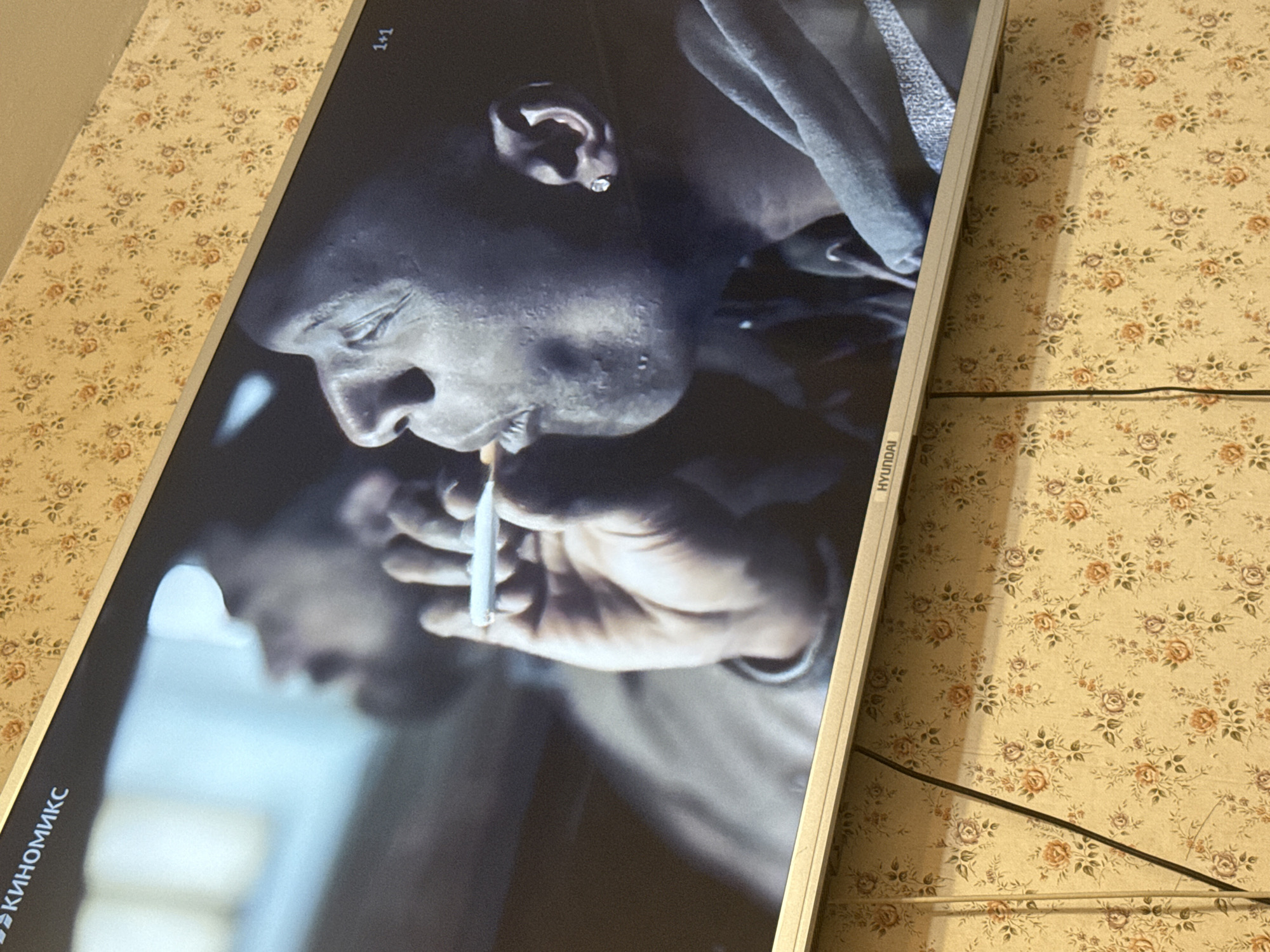 The image shows a close-up of a television screen displaying a black and white image of a person smoking. The screen is surrounded by a floral wallpaper. The television is a Hyundai brand. Text in Cyrillic is visible along the bottom left of the screen and a "1+1" symbol is in the top right corner. There is no information to suggest this image was taken in Mytishchi, Russia.
