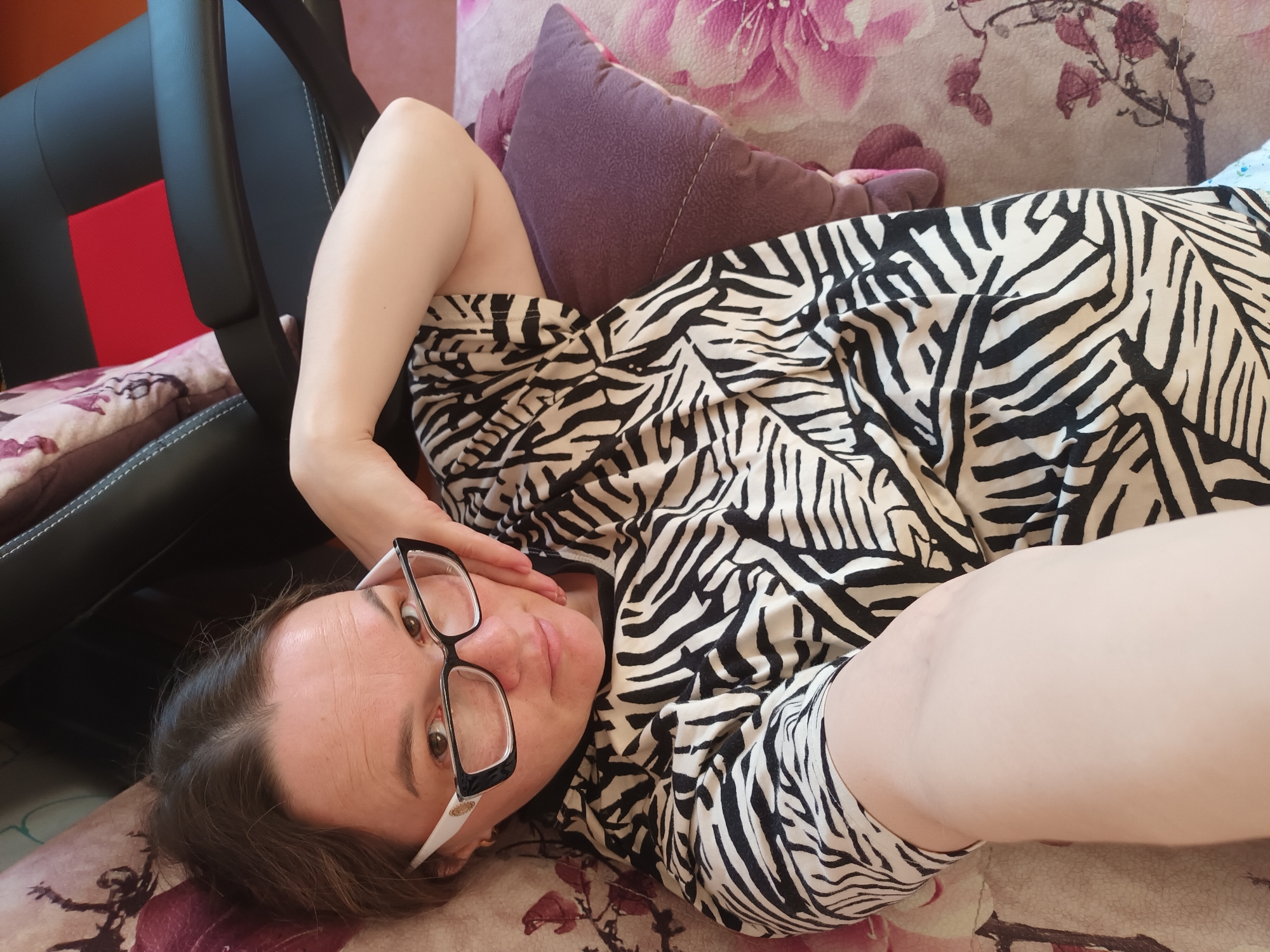 A woman with dark hair and wearing black and white framed glasses lies on a patterned couch, looking directly at the camera. She is dressed in a black and white zebra print top or dress. Her left arm is bent, with her hand resting near her head.
The couch features a light-colored base with a floral or abstract pattern in shades of pink and purple. A dark purple cushion is visible behind her head. In the background, to the left, a black gaming or office chair with a red accent panel is partially visible. A small pink plush toy is also discernible behind the purple cushion.
The setting appears to be indoors, likely a relaxed home environment such as a living room or bedroom, during the daytime. There is no visible text in the image. This scene takes place in Ольховка, Russia.